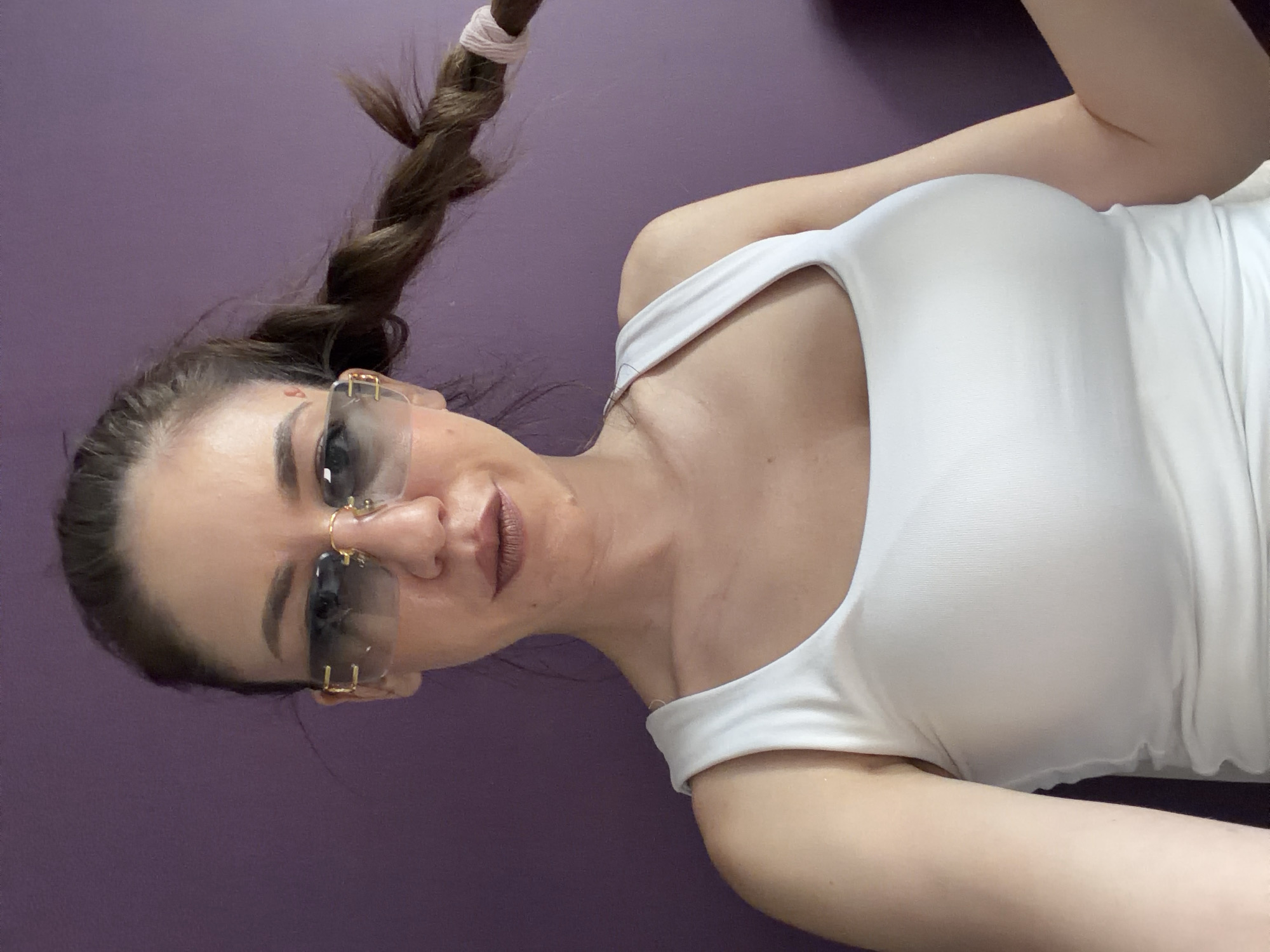 An adult female is depicted in a close-up, selfie-style portrait against a solid, matte purple background. She has dark brown hair, pulled back and styled into a single braid extending over her right shoulder (viewer's right), secured with a light-colored elastic band. She wears large, rimless sunglasses with gold-toned metal accents at the temples and dark gradient lenses. Her eyebrows are dark and well-defined. Her lips are full, coated with reddish-brown lipstick, and slightly parted. A small, faint reddish mark is visible on her left temple (viewer's left). She is wearing a white, form-fitting tank top with a wide, rounded neckline. Her left arm (viewer's right) is slightly raised and bent, with the hand out of frame, suggesting it is holding the camera or phone. Her right arm (viewer's left) is bent, with the hand near the end of her braid. The lighting is soft and even. No other objects or specific environmental markers are discernible.