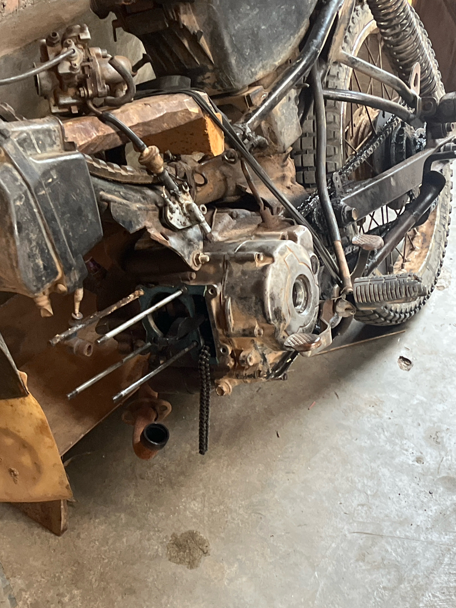 An outdoor, close-up, slightly low-angle, eye-level, eye-level, full shot reveals a severely damaged and rusted motorcycle. The motorcycle is a dirt bike or an older model. The front of the motorcycle is partially visible, including the carburetor. The engine block is exposed, with parts disassembled or missing. A section of the rear wheel and chain is visible. The motorcycle is resting on a concrete surface with some dirt and debris scattered around. The background is out of focus, but appears to be a brick wall or structure. The scene suggests the motorcycle is either abandoned, broken down, or in the process of being repaired or salvaged. The lighting is natural and diffused, indicating it is daytime. The image is from Kebumen, Indonesia.