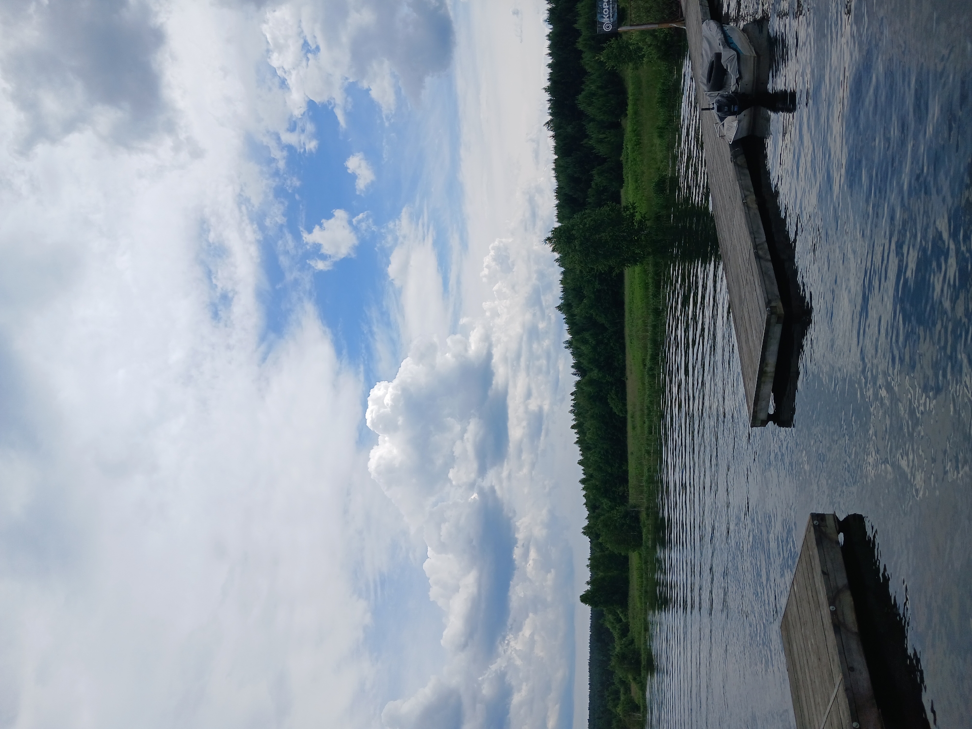 This image shows a body of water with a wooden dock extending into it. A small boat is moored to the dock. In the background, there is a lush green forest and a cloudy sky. The scene appears to be a natural landscape, likely a lake or river, given the presence of the dock and boat. There is a sign on the right side of the dock that appears to have Cyrillic writing on it, indicating it is likely located in Russia. The overall impression is one of tranquility and natural beauty.