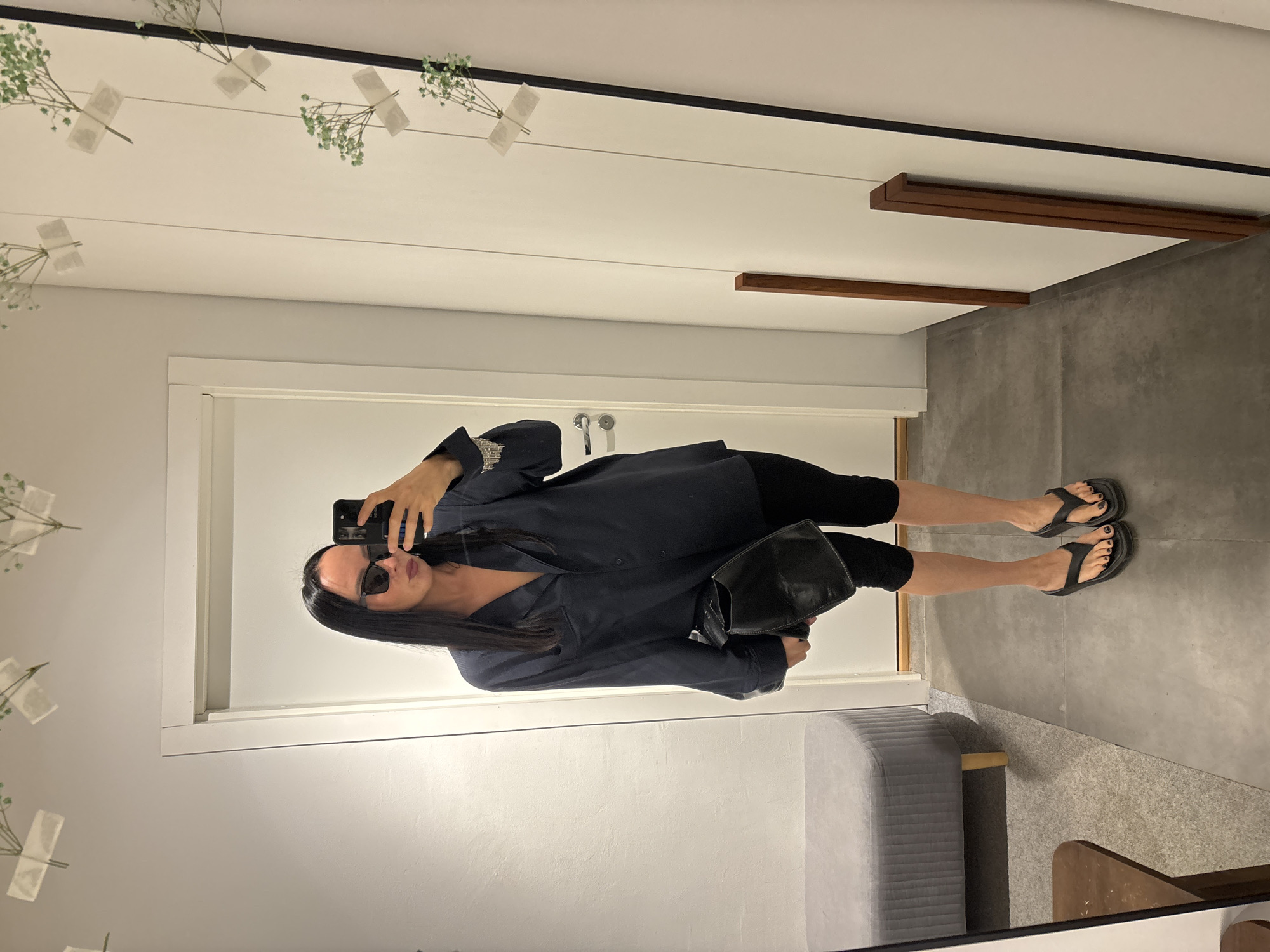 A woman is taking a selfie in what appears to be a fitting room of a clothing store. She is wearing black leggings, a loose-fitting navy blue top, and black sandals. She also has sunglasses on and is holding a black purse. The fitting room is decorated with white walls and minimalist decor, including small floral arrangements attached to the ceiling. There is a gray bench along the bottom of the frame. There is no indication of a city or region in the image.