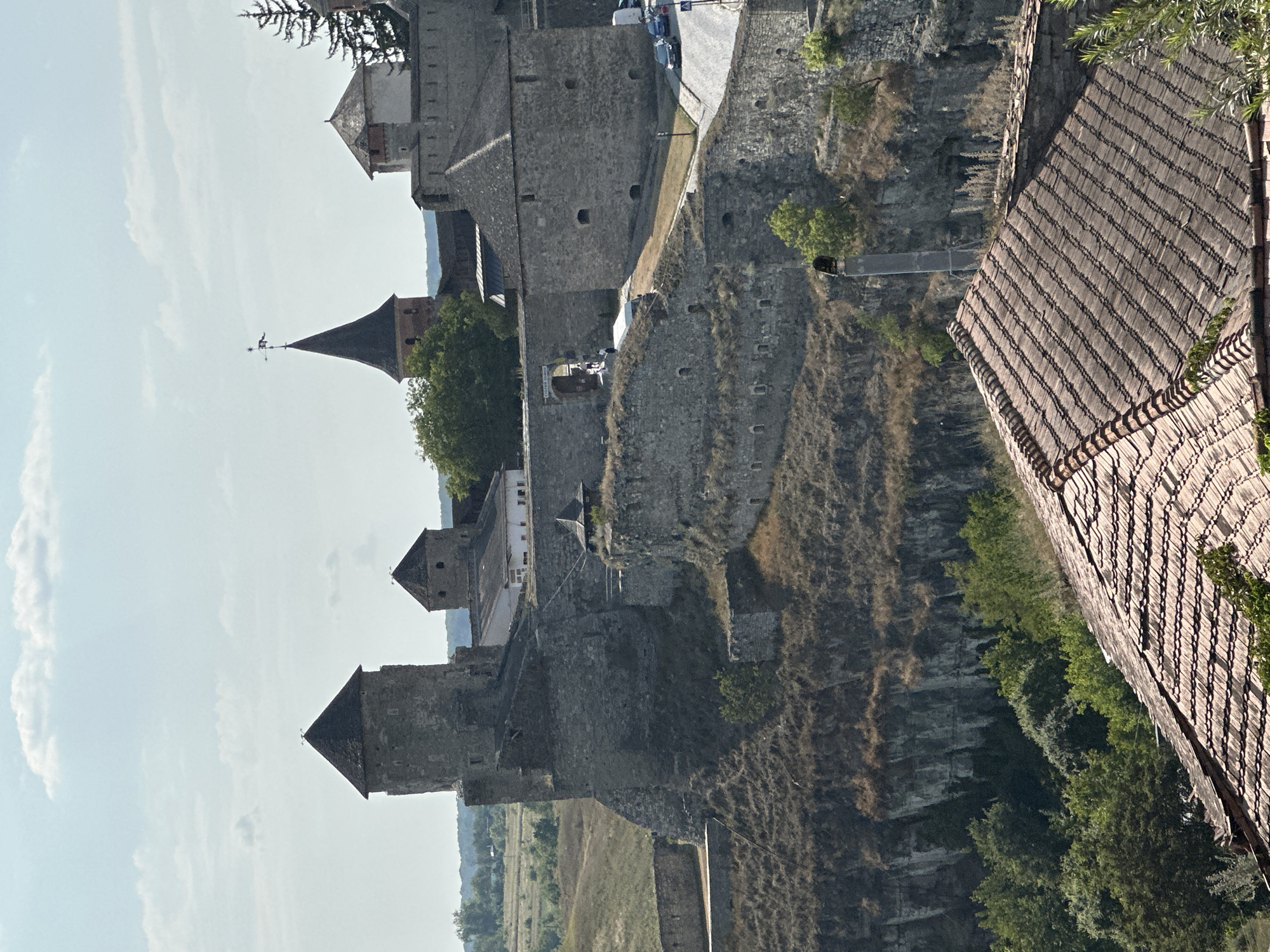 The image captures a panoramic view of the Kamianets-Podilskyi Castle in Ukraine, a historic fortress complex. The castle, built primarily of stone, features several towers of varying shapes and sizes, with conical roofs. A prominent tower on the left has a dark, steeply pitched roof. To its right, another smaller tower is visible. In the background, the castle walls are substantial, with battlements and arched windows. A taller, pointed spire with a weather vane stands out against the sky. The castle is situated on a rocky promontory, with steep cliffs dropping away on the left side, covered in dry grass and sparse vegetation. The foreground shows a tiled roof of a building, with greenery surrounding it. In the middle ground, a road with a few cars is visible, suggesting it's a functioning area within or near the castle. The sky is partly cloudy, with white clouds scattered against a pale blue backdrop. The overall impression is one of a majestic and ancient fortification set within a rugged landscape.