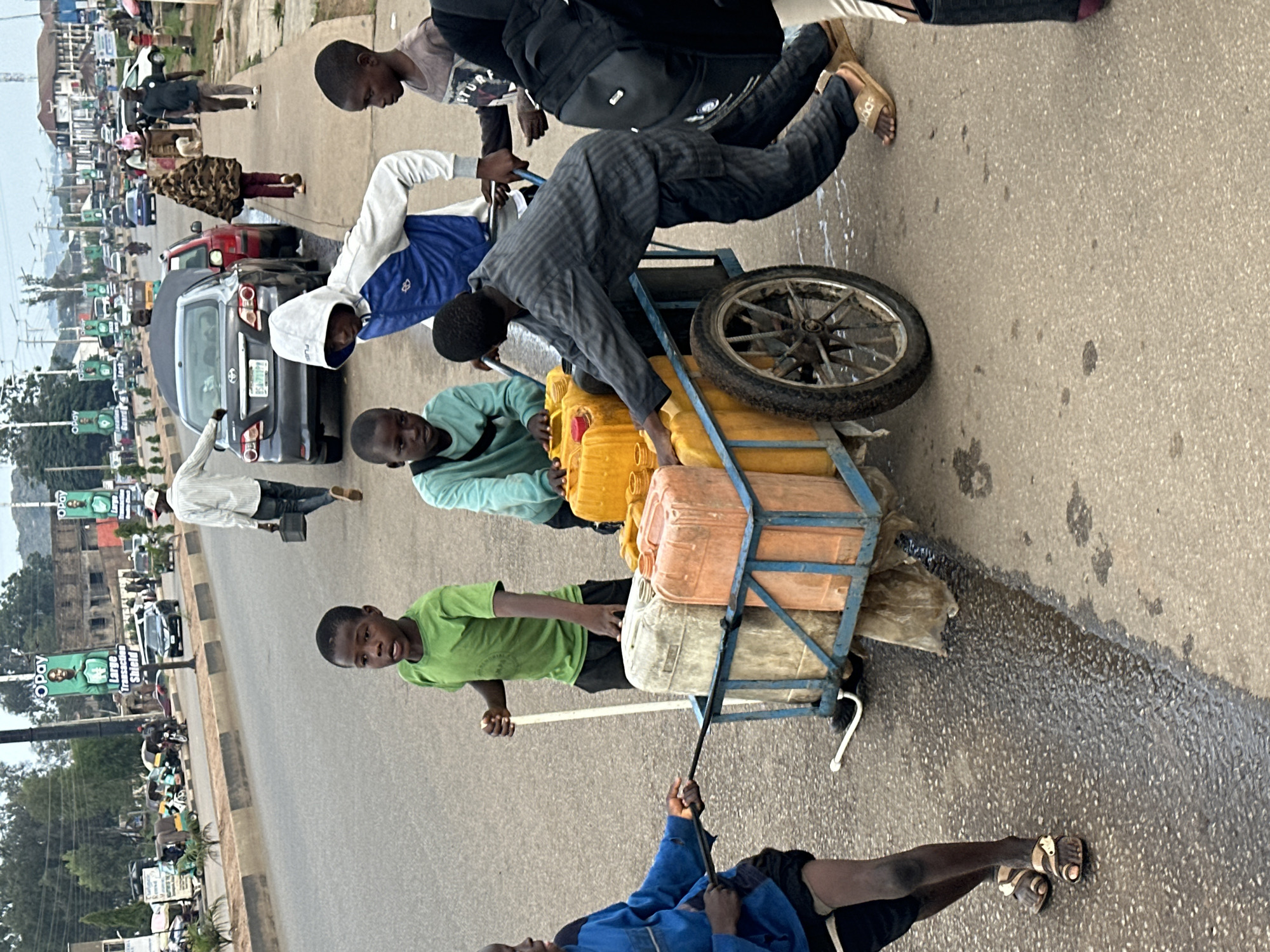 In Jos, Nigeria, a bustling street scene unfolds with several individuals engaged in transporting water. The primary focus is on a multi-tiered cart laden with numerous yellow and orange plastic water containers. Two young boys are actively involved with the cart. One, wearing a green t-shirt, appears to be pushing or guiding it, while another, dressed in a blue jacket, is seen pulling the cart with a rope.
Several other people are present on the street, suggesting a busy urban environment. Among them, a man in a dark striped shirt is leaning over the cart, possibly assisting or supervising. Another individual, wearing a white hooded sweatshirt, stands near the cart, while others are further down the road. The presence of cars and motorcycles indicates ongoing traffic.
The setting appears to be a roadside, with a paved road and a sidewalk or verge. The time of day seems to be daytime, with natural light illuminating the scene. The overall atmosphere is one of activity and movement, typical of a commercial or transport hub. No specific weather conditions are evident, but the wet patches on the road might suggest recent rain or spillage.
Visible text includes "O Pay" on advertisements displayed on poles along the road, a common mobile payment service. The image captures a moment of collective effort and commerce, highlighting the daily lives and activities of the people in this Nigerian city.