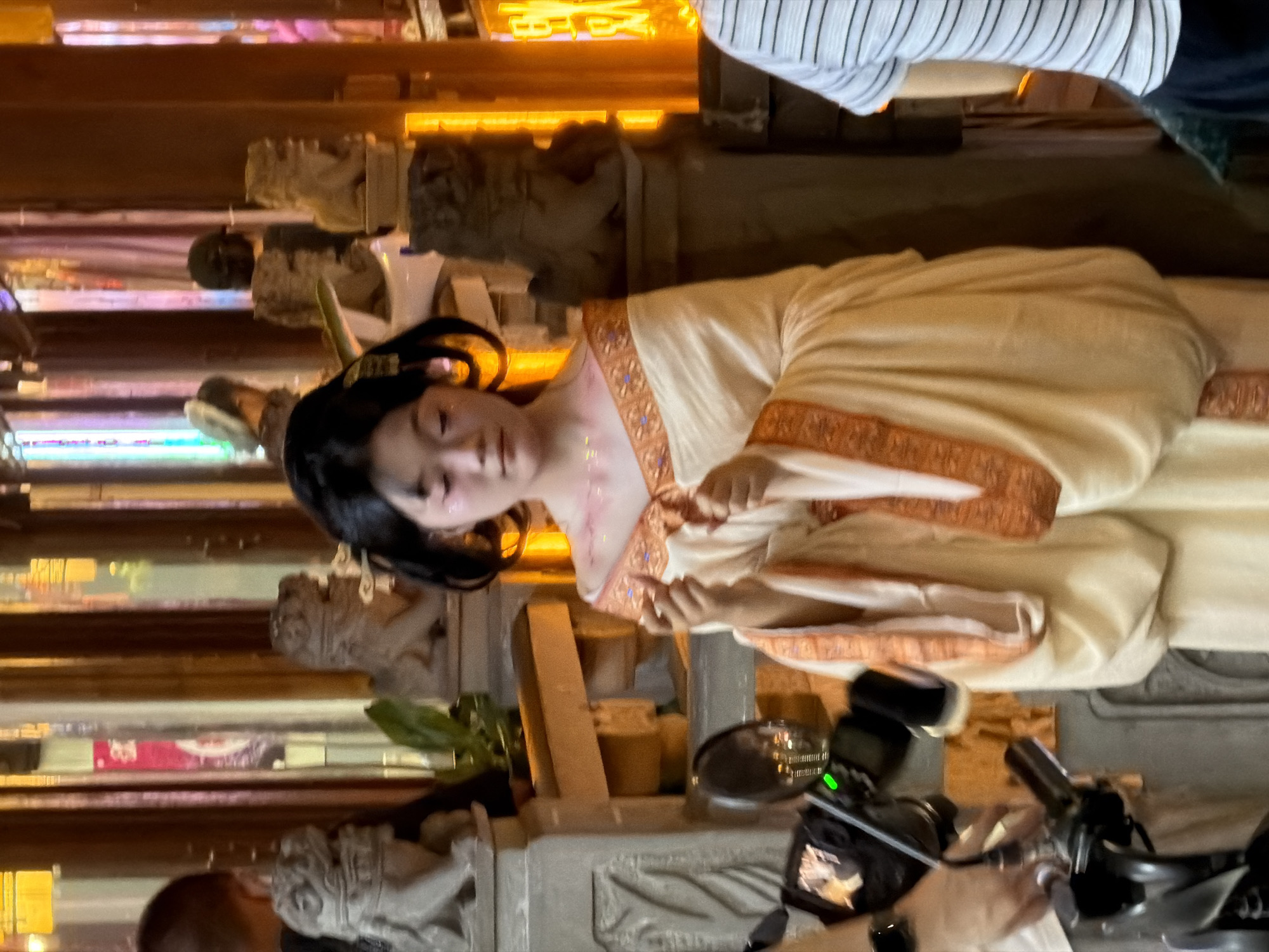 In Yuzhong District, China, a young woman dressed in traditional Chinese attire stands near a scooter, her hair styled in an elaborate updo. She wears a pale cream-colored robe with ornate orange trim around the neckline and sleeves. Her makeup includes subtle pink blush and glitter on her cheeks and shoulders. In her hands, she is performing a gesture with her fingers. The background suggests an indoor setting with elements of traditional Chinese architecture, including carved lion statues, wooden structures, and decorative lighting that casts a warm glow. Part of a person wearing a striped shirt is visible on the right side of the frame.