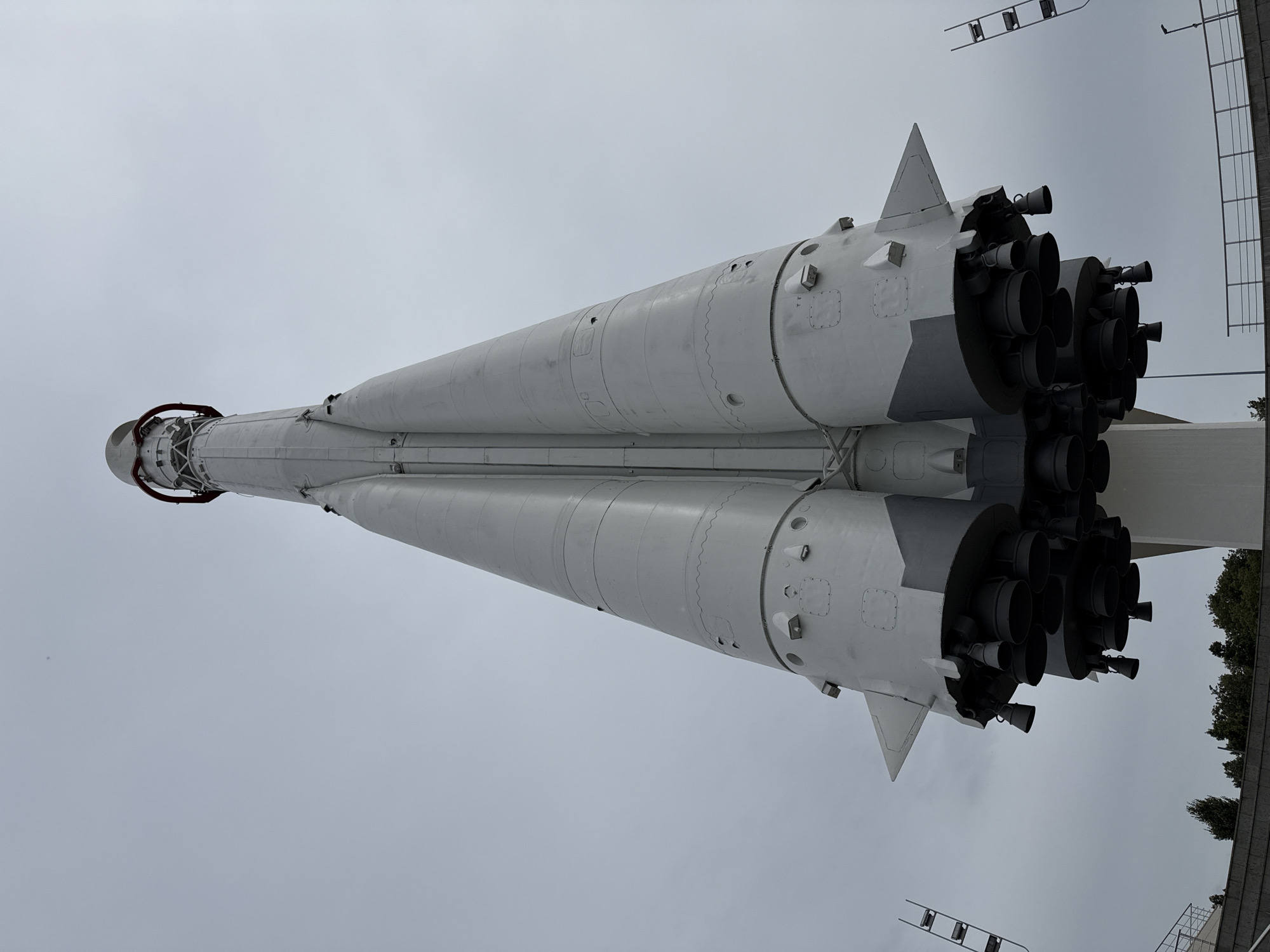 In the city of Kaluga, Russia, a Soviet-era Vostok rocket stands vertically, mounted on a white pedestal. The rocket, primarily painted gray, exhibits its characteristic multi-stage design with multiple booster rockets clustered at the base. The sky above is overcast and pale gray. In the foreground, a low-lying metal fence and some dark green trees are visible, with parts of what appears to be a museum exhibit or viewing area along the bottom edge of the frame, including a set of vertical ladders on the left and right sides. The perspective is low-angle, looking up at the imposing structure.