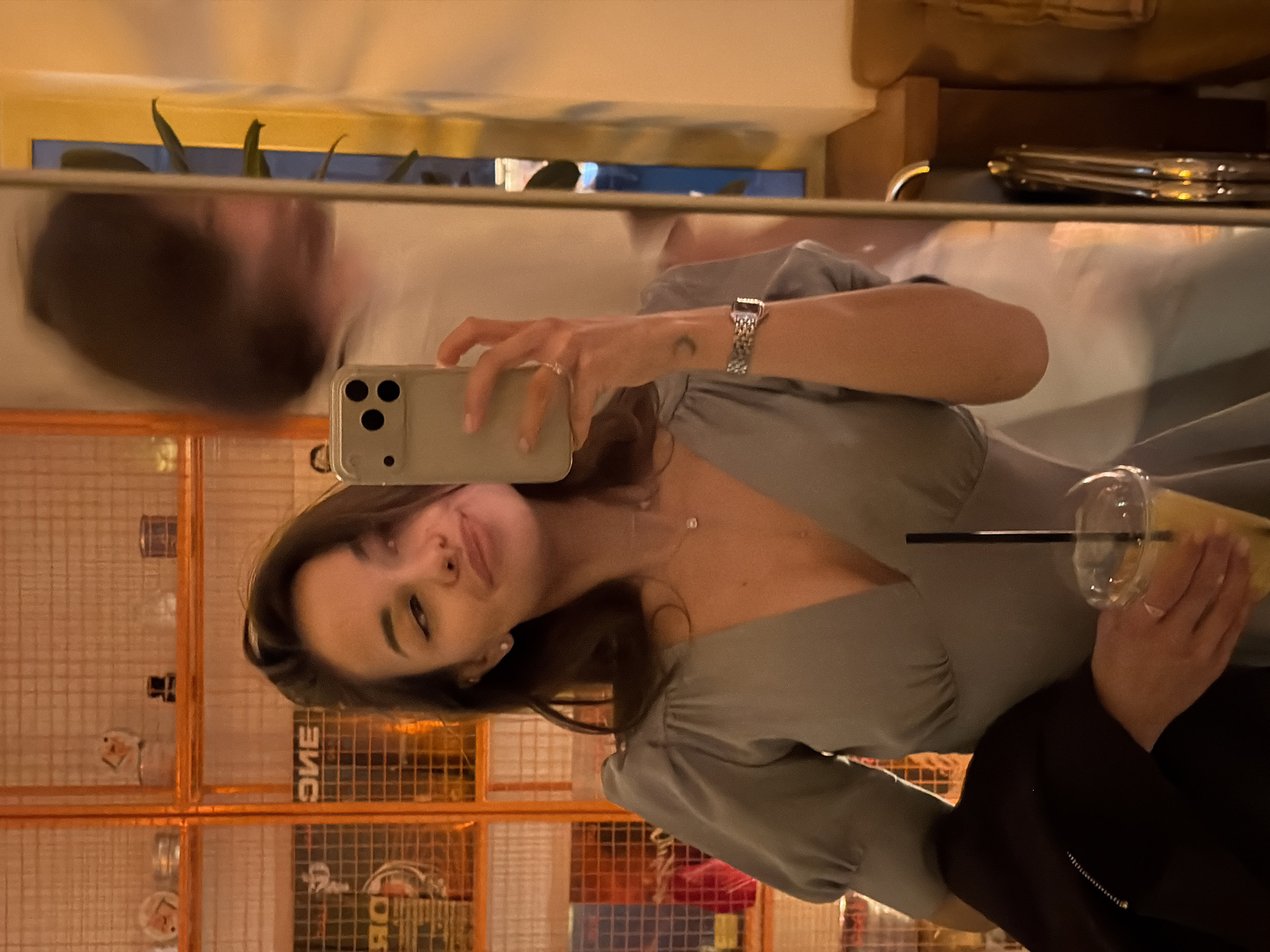 The image shows a selfie taken in a mirror. A young woman with dark hair and fair skin is holding a cell phone, presumably to take the photo. She wears a light-colored dress with puff sleeves and a silver watch. A drink in a clear plastic cup with a straw is also visible in her hand.
In the mirror's reflection, a man with dark hair can be seen.
The background reveals an interior setting with shelves or racks behind the woman, possibly a store or cafe with orange walls and patterned metal screens. The reflection in the mirror suggests a city setting, which could be Krasnoyarsk, Russia. More details on precise location can't be determined.