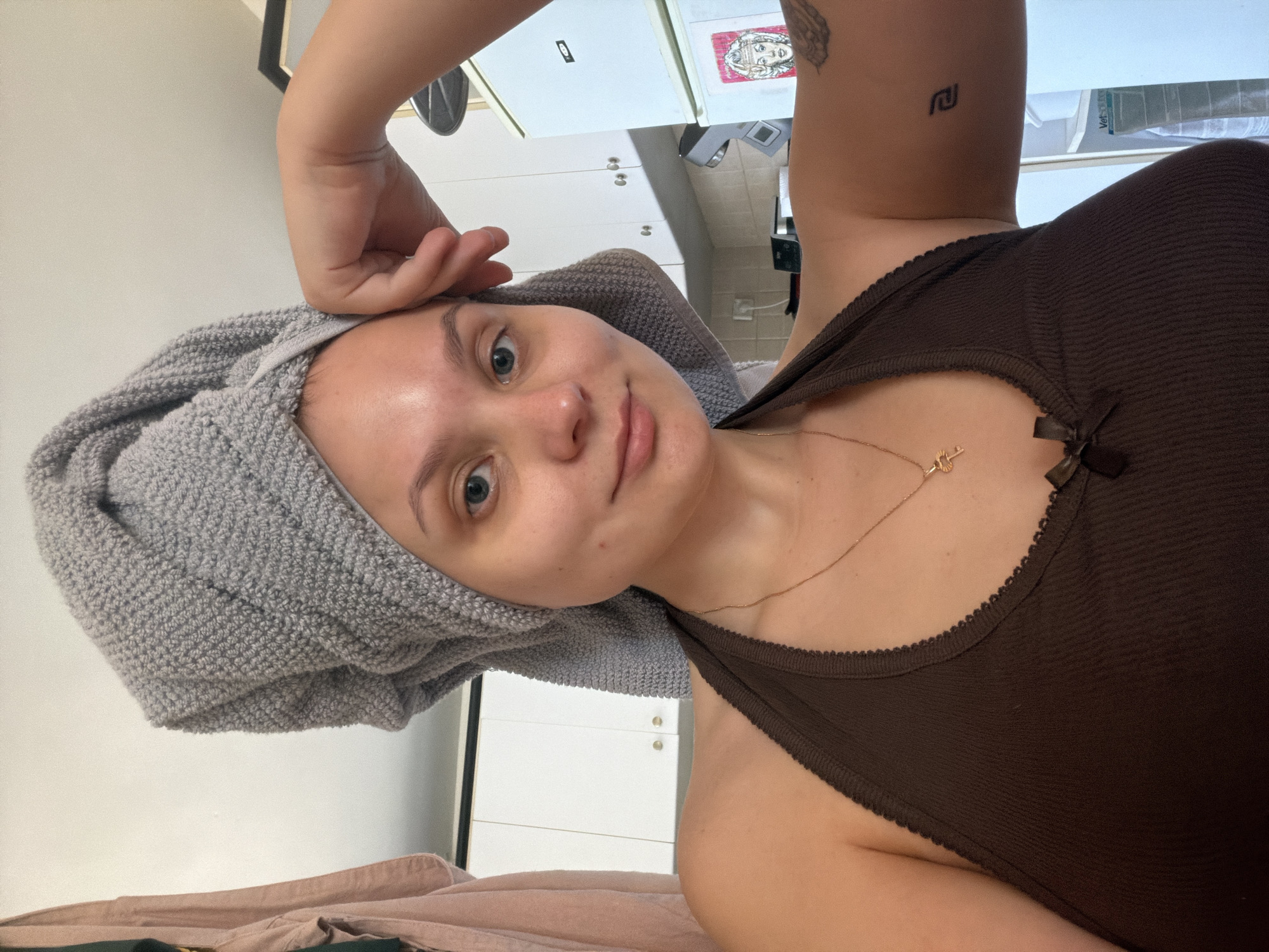 A selfie-style, indoor shot of a fair-skinned individual. The person has a gray towel wrapped around their head, covering their hair. They are wearing a dark brown tank top with a bow detail. A thin gold necklace with a charm is visible around their neck. The person's right arm is raised, with their hand near their forehead. There are two visible tattoos: one on the right forearm and another above it. The background appears to be a kitchen setting with white cabinets and appliances. It is not possible to determine if the location is in Tel-Aviv, Israel from the image alone.