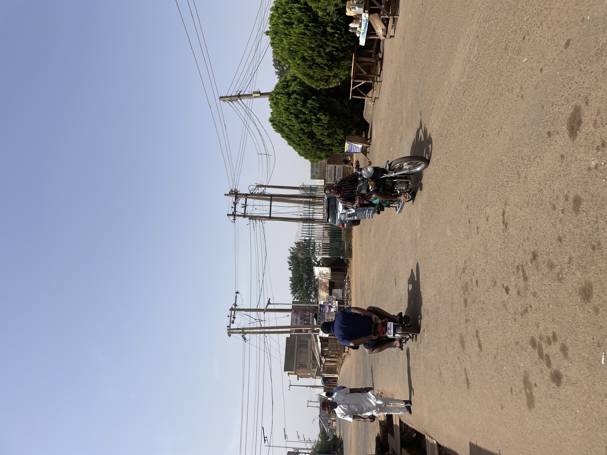 The image depicts a street scene in Jos, Nigeria. The view is from ground level, looking along a paved road. Three people are visible: two men are riding motorcycles in the same direction along the road, and one man is walking in the same direction. The man on the first motorcycle is wearing a striped shirt and has a box strapped to the front of his bike. The man on the second motorcycle is wearing a blue shirt. The walking man is wearing a white outfit. Power lines run along the side of the road, supported by wooden poles. Buildings and vegetation line the street. The sky is clear.