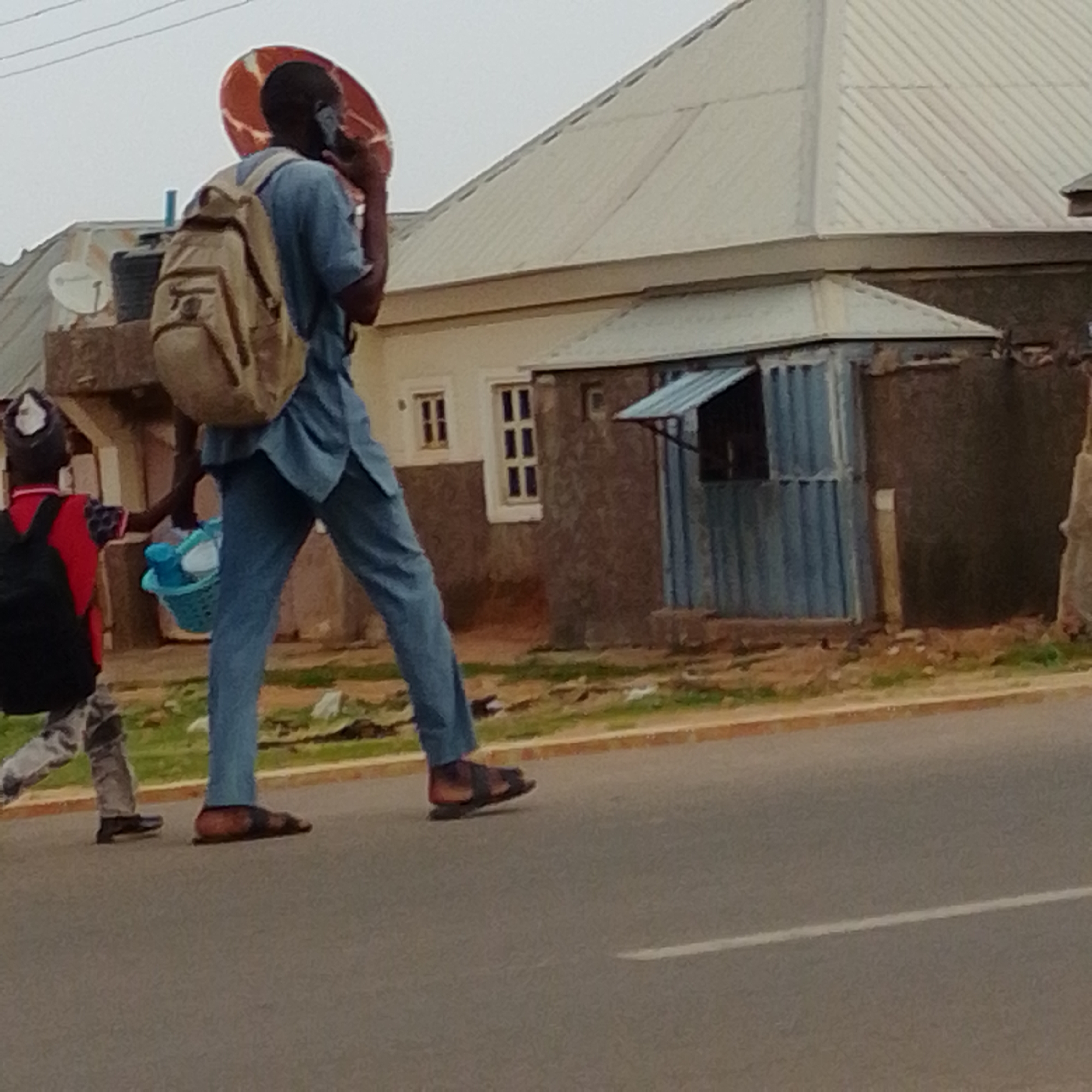 Two individuals are walking on a paved road in Gombe, Nigeria. The person in the foreground, facing away from the camera, is wearing a blue long-sleeved shirt and pants, a light brown backpack, and dark sandals. They are holding a mobile phone to their ear and have a large, round, reddish-orange object strapped to their back, partially obscured by the backpack. This object appears to be a satellite dish.
To the left, a younger person is walking in the same direction, also facing away from the camera. They are wearing a red shirt, camouflage patterned pants, and dark shoes, and carrying a black backpack. They are holding a light blue basket filled with white and blue items.
The scene appears to be set during the daytime, with overcast or cloudy weather indicated by the diffused light. In the background, there are buildings with corrugated metal roofs and a small, blue structure that resembles a kiosk or stall. The road has a white line marking its center. The overall setting suggests a residential or commercial area in a Nigerian town. No text is visible in the image.