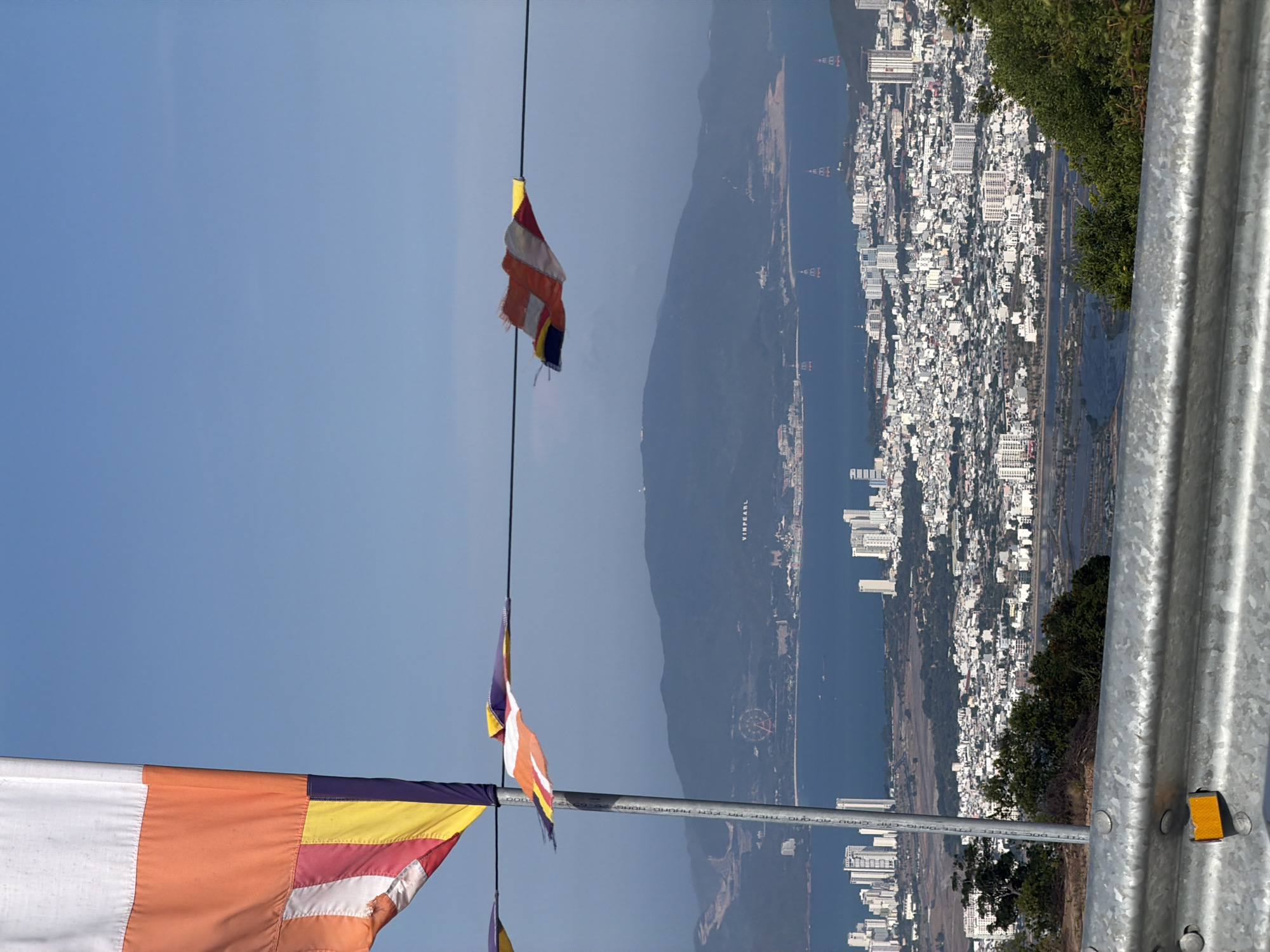 A panoramic view of Nam Nha Trang Ward, Vietnam, is captured from an elevated vantage point. The urban landscape is dominated by densely packed white buildings, extending towards a body of water that reflects the sky. Mountains rise in the background, creating a natural contrast with the built environment. In the foreground, a metal railing runs horizontally. A series of Buddhist flags hang in the sky, adding color and texture to the scene.