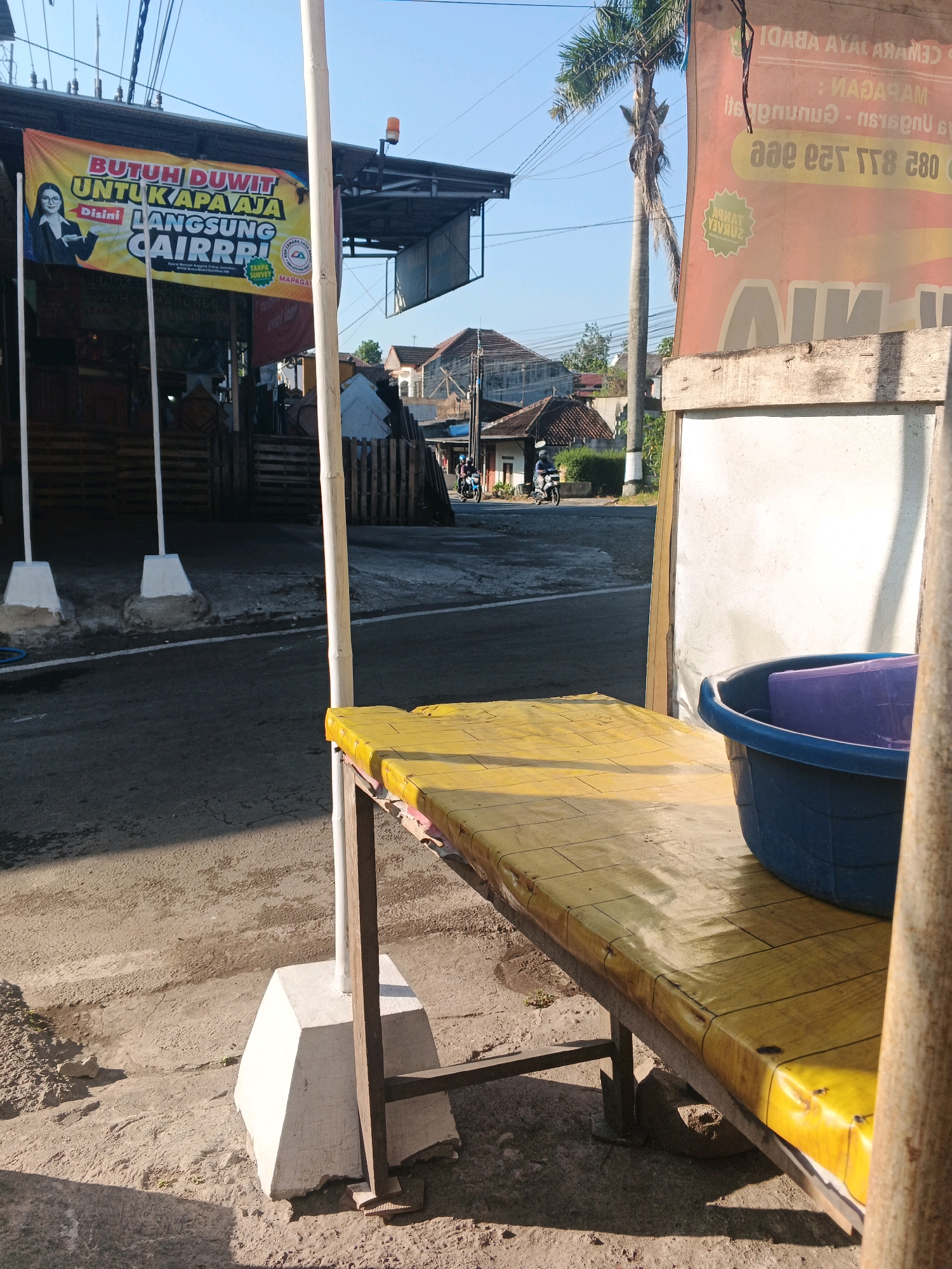 The image captures a sun-drenched street scene in the city of Semarang, Indonesia. In the foreground, a weathered yellow bench with a rough texture sits on a concrete and dirt ground. Beside it, a blue plastic basin contains a purple object. A white pole stands vertically in front of the bench, casting a shadow. In the background, a large banner with Indonesian text and an image of a woman is displayed on a building. Further down the street, houses and trees are visible under a clear blue sky. The overall atmosphere is that of a typical urban street in Indonesia on a bright day.