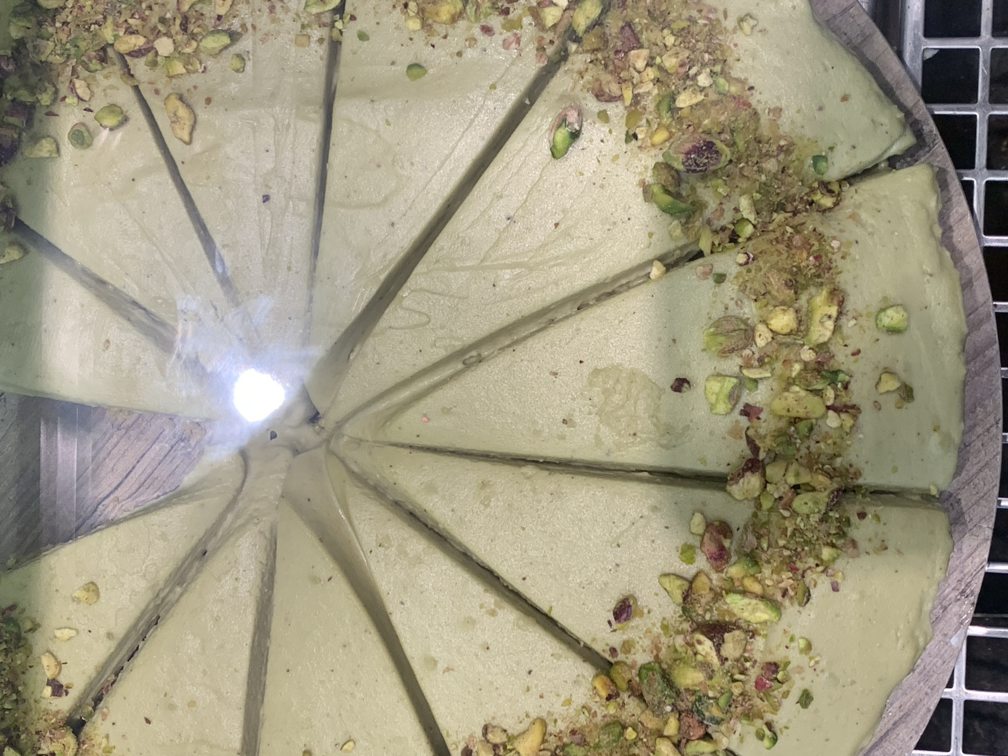 The image shows a circular pistachio cake that has been pre-sliced into approximately 10 pieces. The cake has a pale green hue and is topped with chopped pistachios, predominantly around the outer edges of the slices. The cake sits on a round, wooden base. The background appears to be a stainless-steel surface, possibly a refrigerated display case. The bright light suggests the cake is being viewed through some transparent barrier, likely glass or plastic, indicating that it is on display. This cake could be located at a bakery or patisserie in Barcelona.