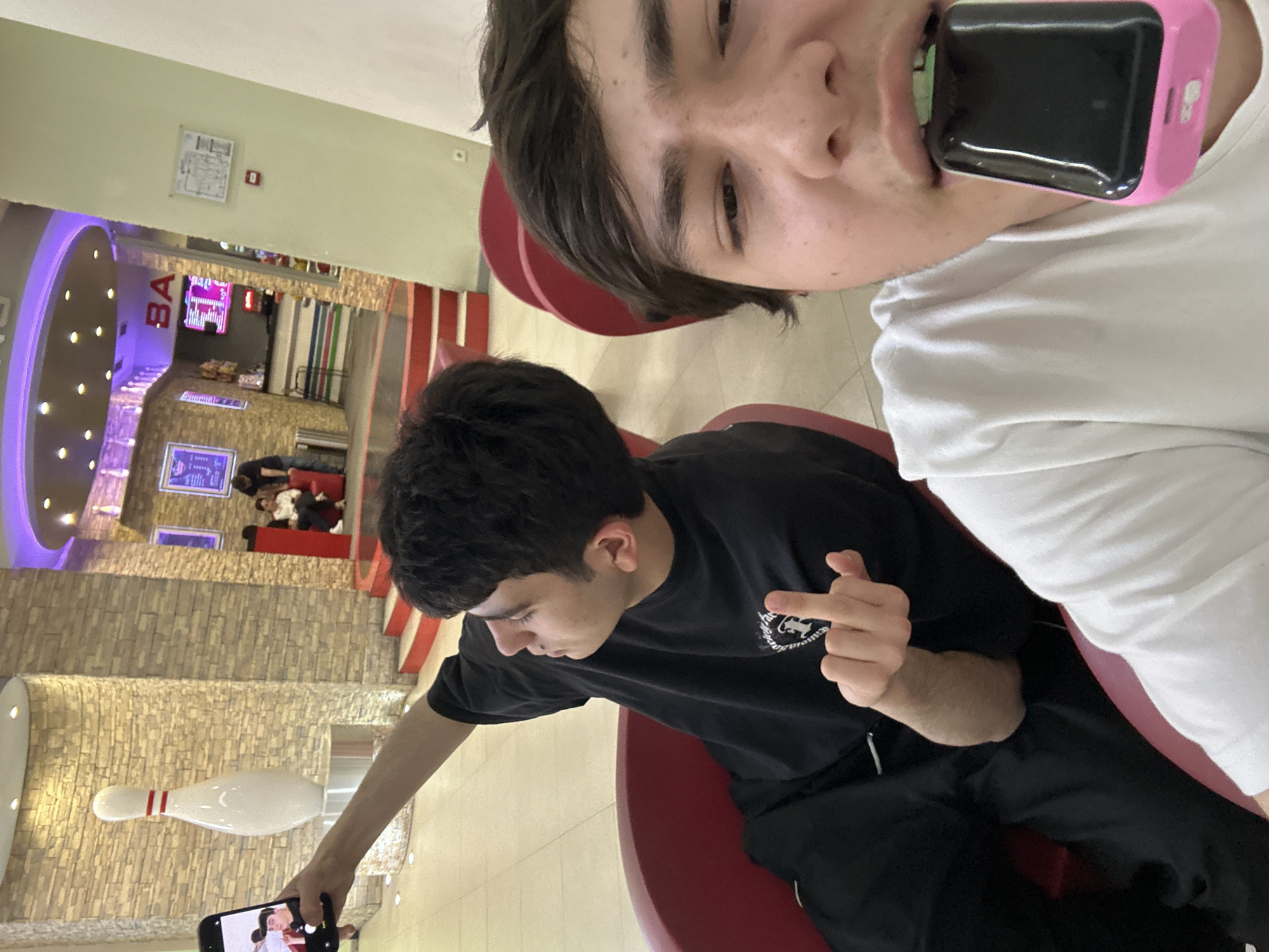 Two young men are seated in red chairs in a bowling alley, taking selfies. One man holds a phone in his mouth and wears a white t-shirt. The other man, wearing a black t-shirt and black pants, holds a phone up to take a photo, while simultaneously extending his middle finger. A bowling pin can be seen in the background. The environment includes the bowling alley's interior, with its stone-patterned wall and bowling lanes visible in the distance. The location is Unknown, Unknown.