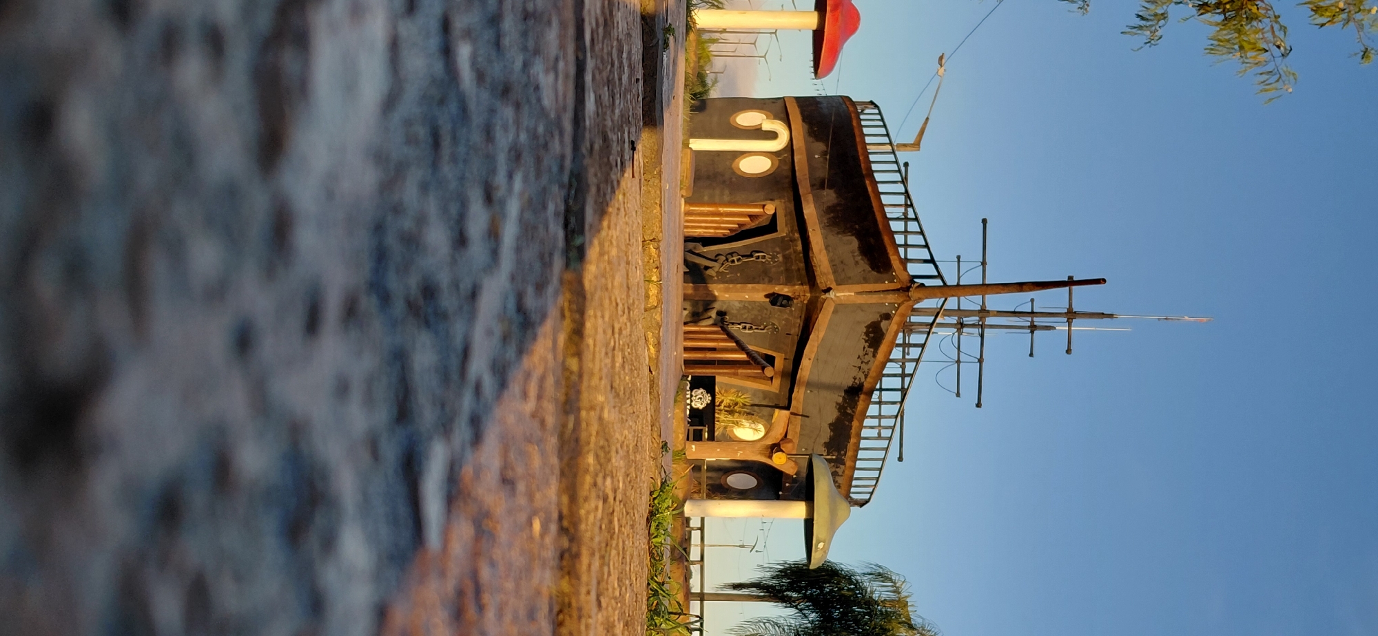 This is a low-angle, full-shot photograph of a whimsical, ship-shaped structure situated outdoors in the city of Encarnación, Paraguay. The structure appears to be a building designed to resemble the bow of a dark-colored ship, complete with a prominent mast and rigging. The foreground is dominated by a rough-textured, stone-like surface, casting a long shadow to the left, indicating the sun is low in the sky, likely during sunrise or sunset.
To the left of the ship structure, a tall, red mushroom-shaped sculpture is visible, and to the right, a similar green mushroom-shaped structure stands. The background reveals a clear blue sky and some distant trees and streetlights, suggesting an urban or park-like setting. The ship structure has multiple round windows and wooden doors, and decorative anchors and chains are attached to its facade. The overall scene evokes a playful and imaginative atmosphere, characteristic of a themed attraction or public art installation.