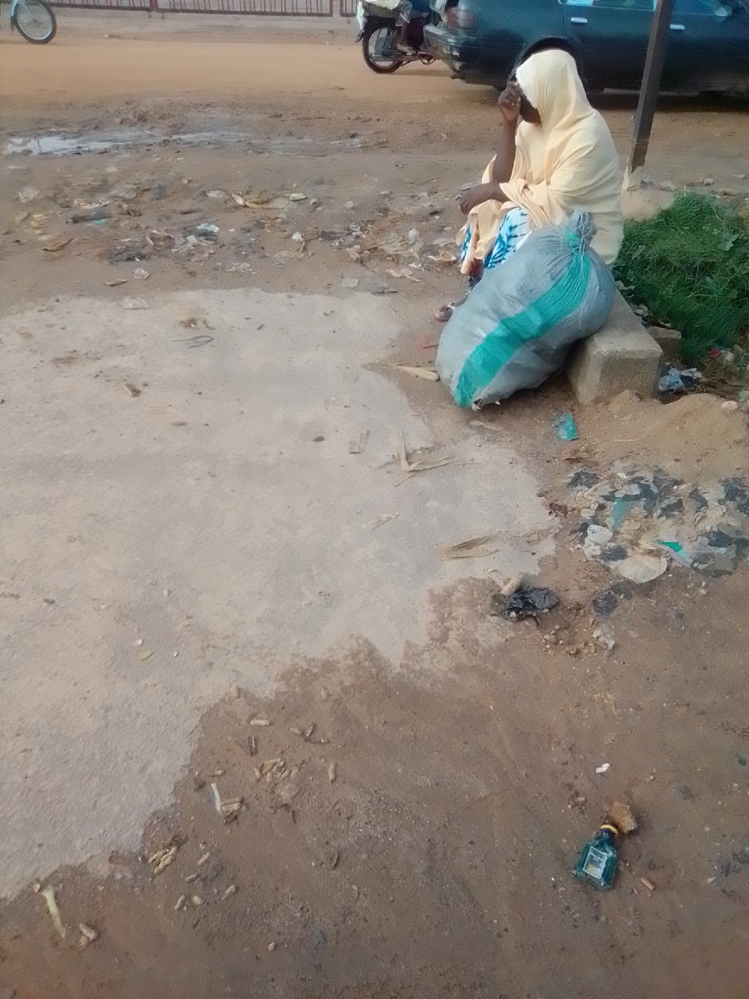 The image captures a street scene in Gombe, Nigeria. A person, draped in a light yellow headscarf and patterned garment, is seated on a concrete block outdoors. Beside them, a large grey trash bag with a green stripe is placed on the ground. The surrounding environment is unkempt, with scattered debris, including plastic waste, visible on the dirt and concrete surfaces. A dark-colored vehicle and parts of a motorcycle are partially visible in the background, suggesting a public thoroughfare. The ground shows evidence of recent moisture, with puddles and damp patches. A small green glass bottle lies on the ground near the lower right corner of the frame. The overall impression is one of a less-than-pristine urban setting.