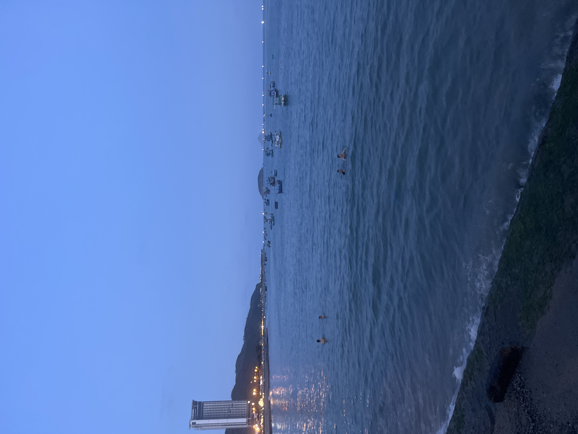 The image depicts the shoreline of Bac Nha Trang Ward, Vietnam at dusk. The sky is a uniform blue, transitioning to a darker hue near the horizon. A body of water, likely the sea, occupies the right half of the frame. Gentle waves break along a shoreline characterized by a dark, moss-covered edge. In the water are two individuals engaged in swimming. Further out to sea, several boats are anchored. Along the shoreline, there is a stretch of land with illuminated streetlights and buildings. The most prominent structure is a tall building on the left with its lights reflecting on the water. A mountain outlines the shoreline.