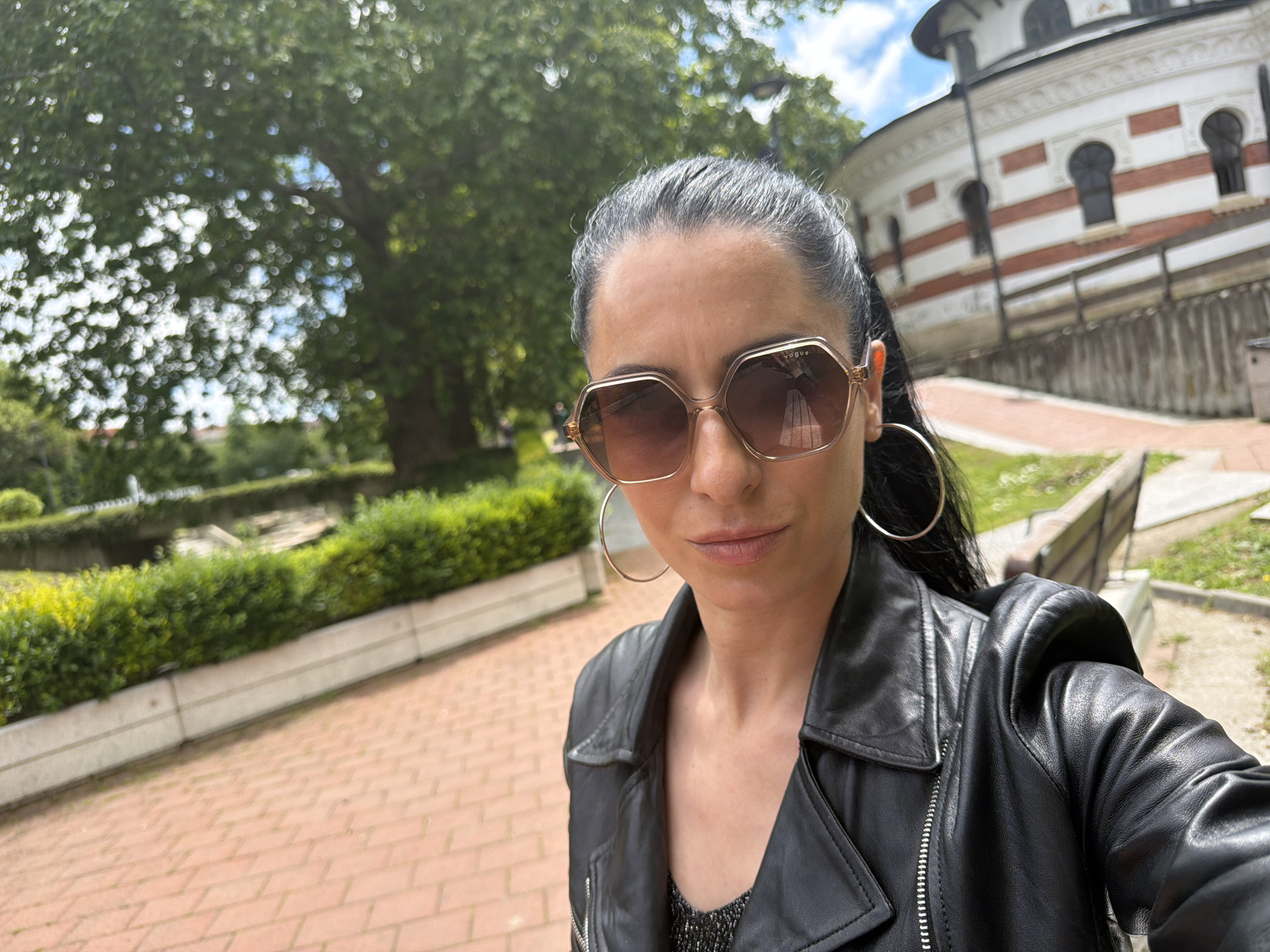 A selfie of a woman wearing large gold hoop earrings and Vogue sunglasses. She has dark hair pulled back and is wearing a black leather jacket. In the background is a brick pathway, greenery, a tree, and a white building with red and brown stripes. Based on the architecture, the building appears to be the Mausoleum Ossuary of Pleven Epopee 1877. The photo was likely taken in the city of Pleven, Bulgaria.