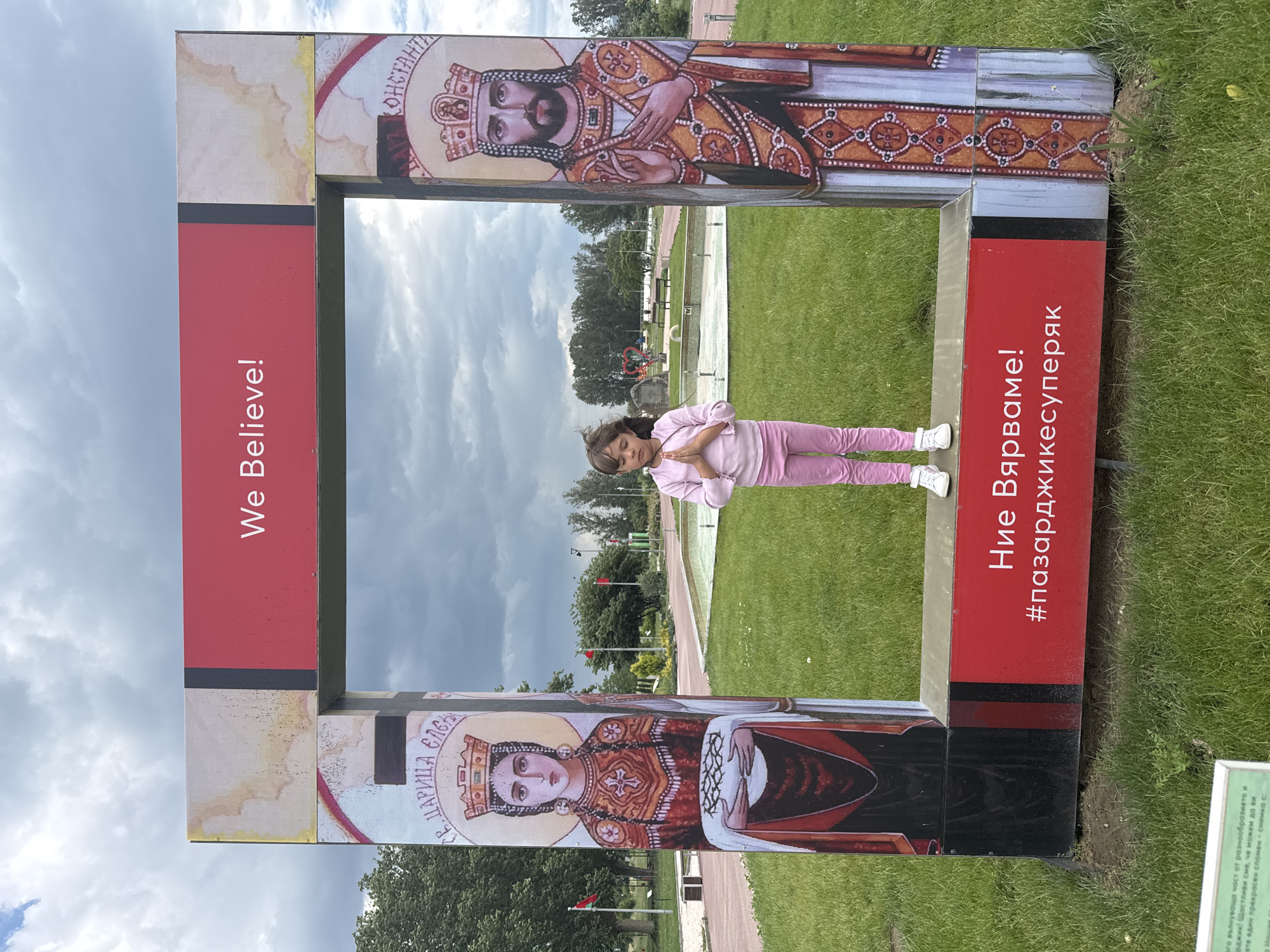 A young girl in a pink outfit stands within a frame-like structure on a grassy field, against a backdrop of a partly cloudy sky. The frame is a rectangular construct, adorned with religious iconography and text. The upper panel of the frame features a depiction of a king-like figure, and the lower panel shows a queen-like figure. The frame also includes sections with red backgrounds and text in what appears to be a non-English language. The girl is positioned centrally within the frame, with her hands folded in front of her, as if in prayer. The background consists of a green lawn, trees, and a walking path, possibly in a park. The OCR text included in the image indicates "We Believe!" and what might be a hashtag "#пазарджикесуперяк". The location is referenced in the OCR as possibly relating to Пазарджик, Bulgaria.
