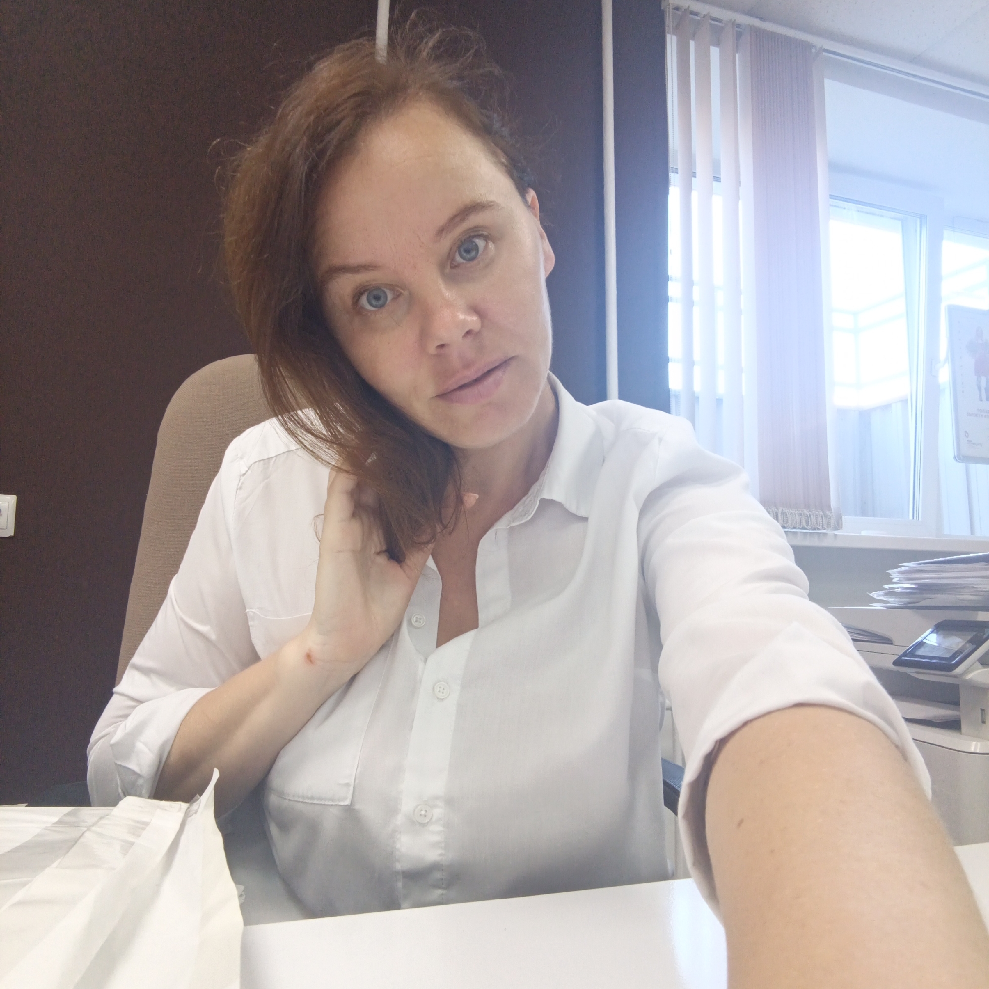 A woman with auburn hair and blue eyes is taking a selfie at a desk in what appears to be an office environment. She is wearing a white button-up shirt. Her left hand is touching her hair, and she has a small red mark on her left wrist. A printer or copier is visible on the desk behind her. Vertical blinds are present on a window in the background. The location is the city of Chaikovsky, Russia.