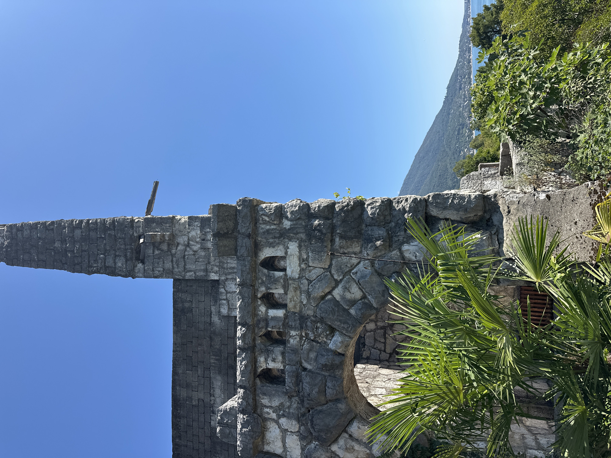 A stone structure, possibly a historical ruin or a defensive fortification, stands against a bright blue sky. The structure is made of large, rough-hewn stones and features an arched opening partially obscured by lush green palm fronds in the foreground. Above the arch, there are several small, recessed openings, resembling crenellations or decorative niches. A tall, slender tower rises from the main structure, topped with broken stone. A wooden beam protrudes from the side of the tower. To the right of the stone structure, a verdant hillside slopes down towards a body of water, likely the Black Sea, with a town or village nestled along the coast and a mountain range in the background. The overall scene suggests a sunny, Mediterranean-like climate. The location is identified as Abaata, Abkhazia.