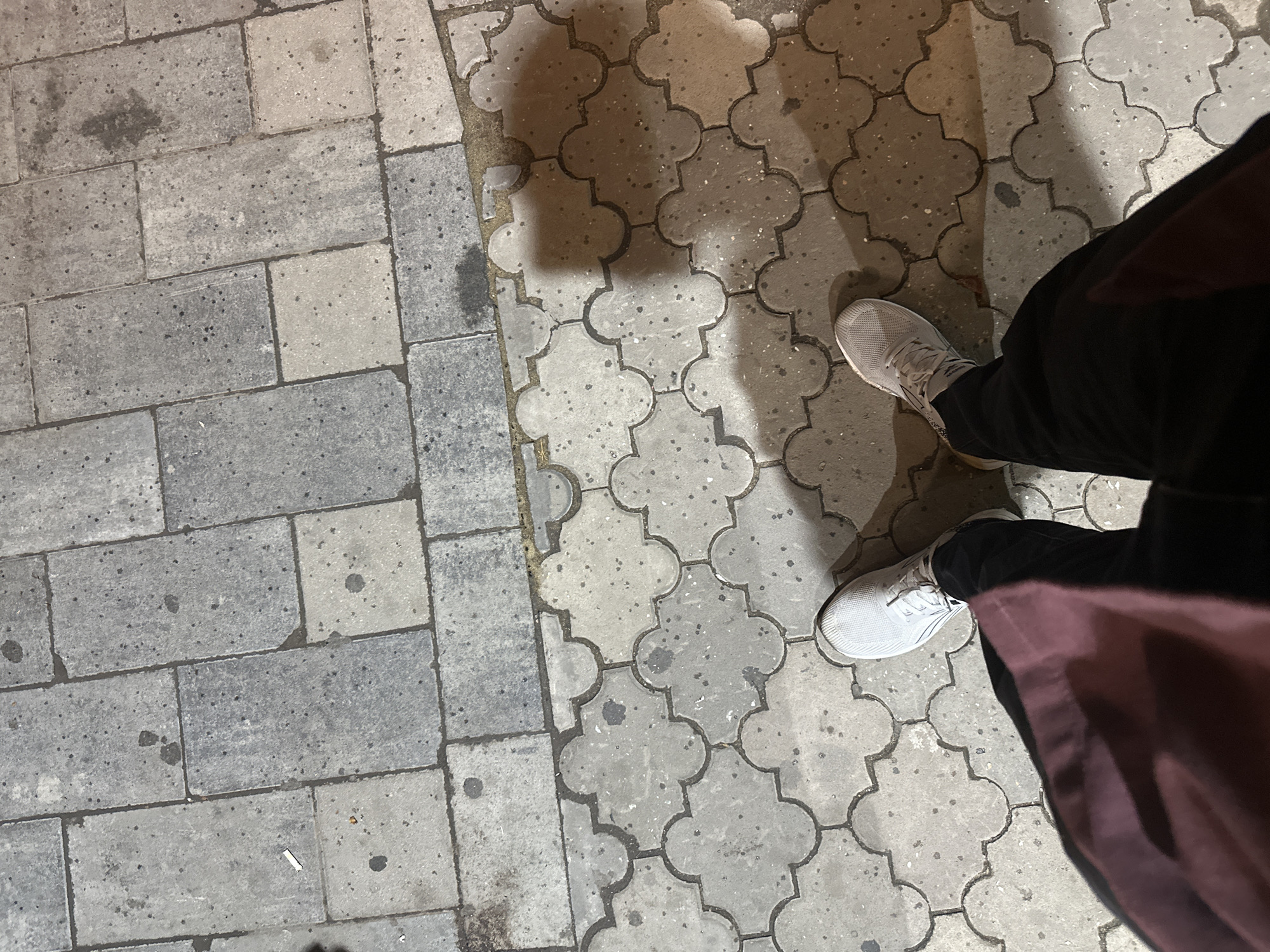 The image shows a first-person perspective of someone standing on a paved sidewalk in the city of Kostanay, Kazakhstan. The sidewalk is composed of two types of paving stones: rectangular grey slabs in the upper portion of the image, and interlocking cloverleaf-shaped grey pavers in the lower portion. Dark spots and some lighter smudges are visible on the pavers, suggesting they are wet or dirty. A discarded cigarette butt is present on the rectangular pavers. The person is wearing black pants and white athletic shoes. A portion of a maroon garment, possibly a jacket or coat, is visible in the lower left corner of the frame. The lighting suggests it is nighttime, and a shadow of the person is cast to the right.