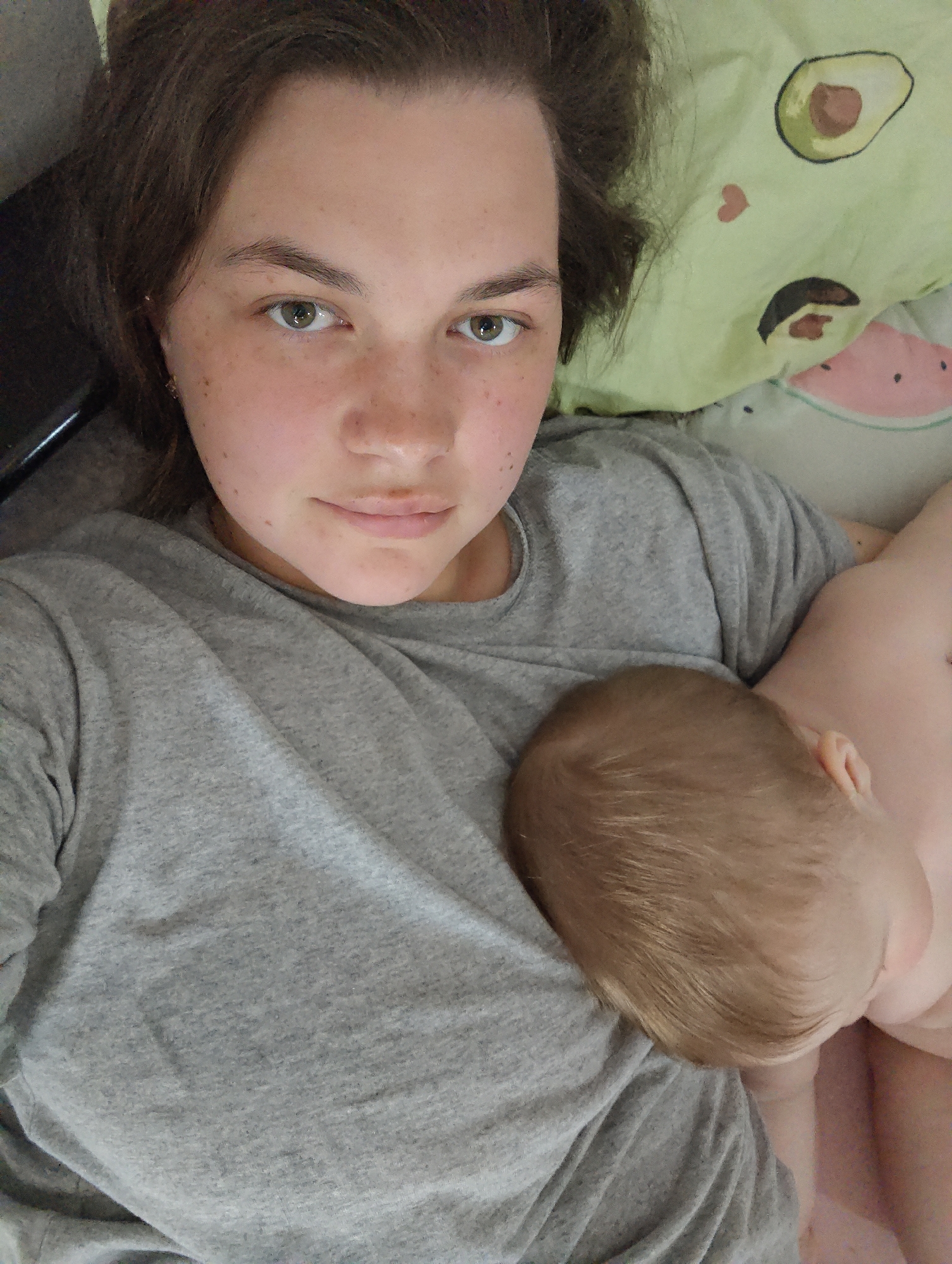 A woman with fair skin, freckles, and brown hair is lying down in a bed, looking at the camera. She is wearing a gray t-shirt. A baby with fair skin and light brown hair is lying on her chest. The bedding includes a pillow with avocado and watermelon designs. There are no features indicating the location being in Chaikovsky, Russia.