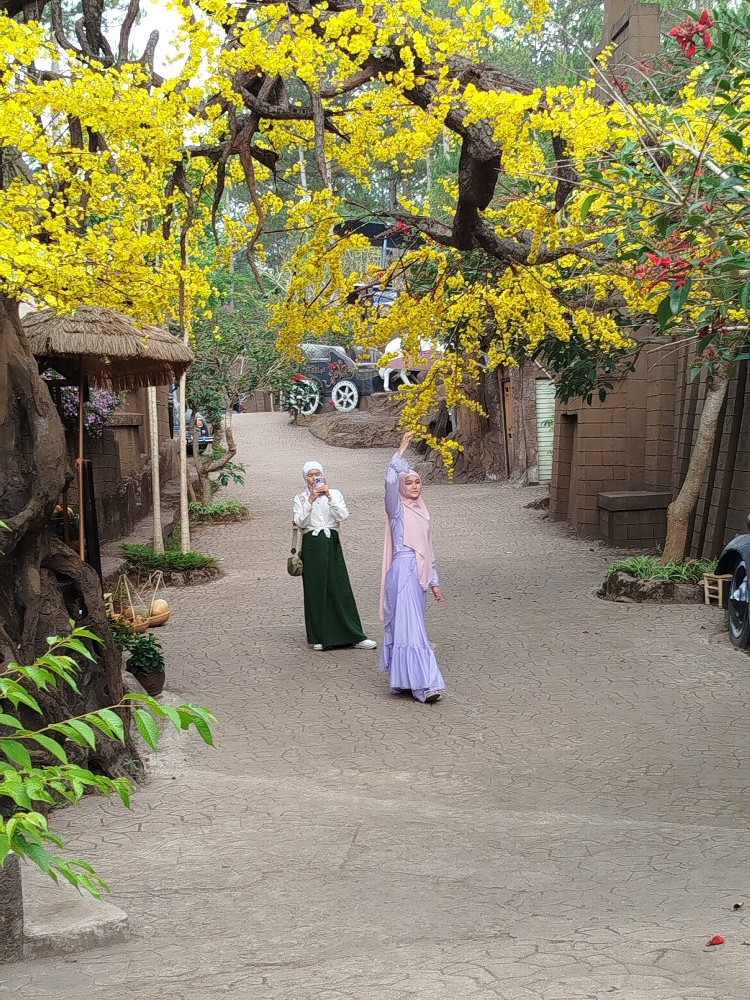 The image captures a scene in Da Lat, Vietnam, showing two women dressed in modest clothing and headscarves on a stone-paved path. The woman on the left is taking a photo with her phone, while the woman on the right has her arm raised towards a tree with yellow flowers. In the background, there is a decorative carriage, buildings with thatched roofs and stone-like facades, and various trees and greenery. The overall environment suggests a tourist attraction or a themed area within Da Lat.