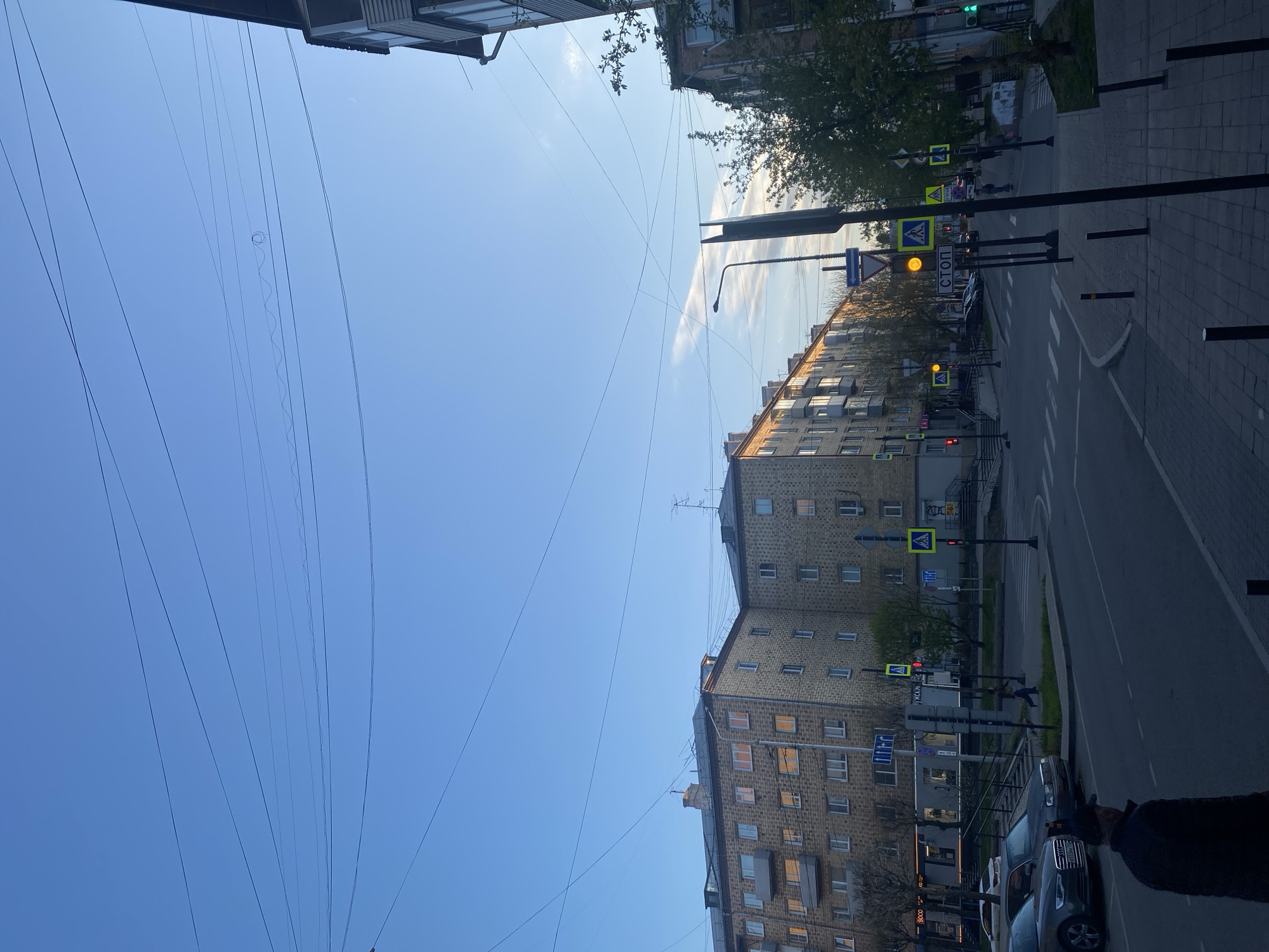 The image depicts a street scene in Krasnoyarsk, Russia. It shows a multi-story brick building with several windows, a street with pedestrian crosswalks, and various traffic signs. Power lines stretch across the sky. Cars are parked along the street. A traffic light shows a yellow light. A few pedestrians are present.