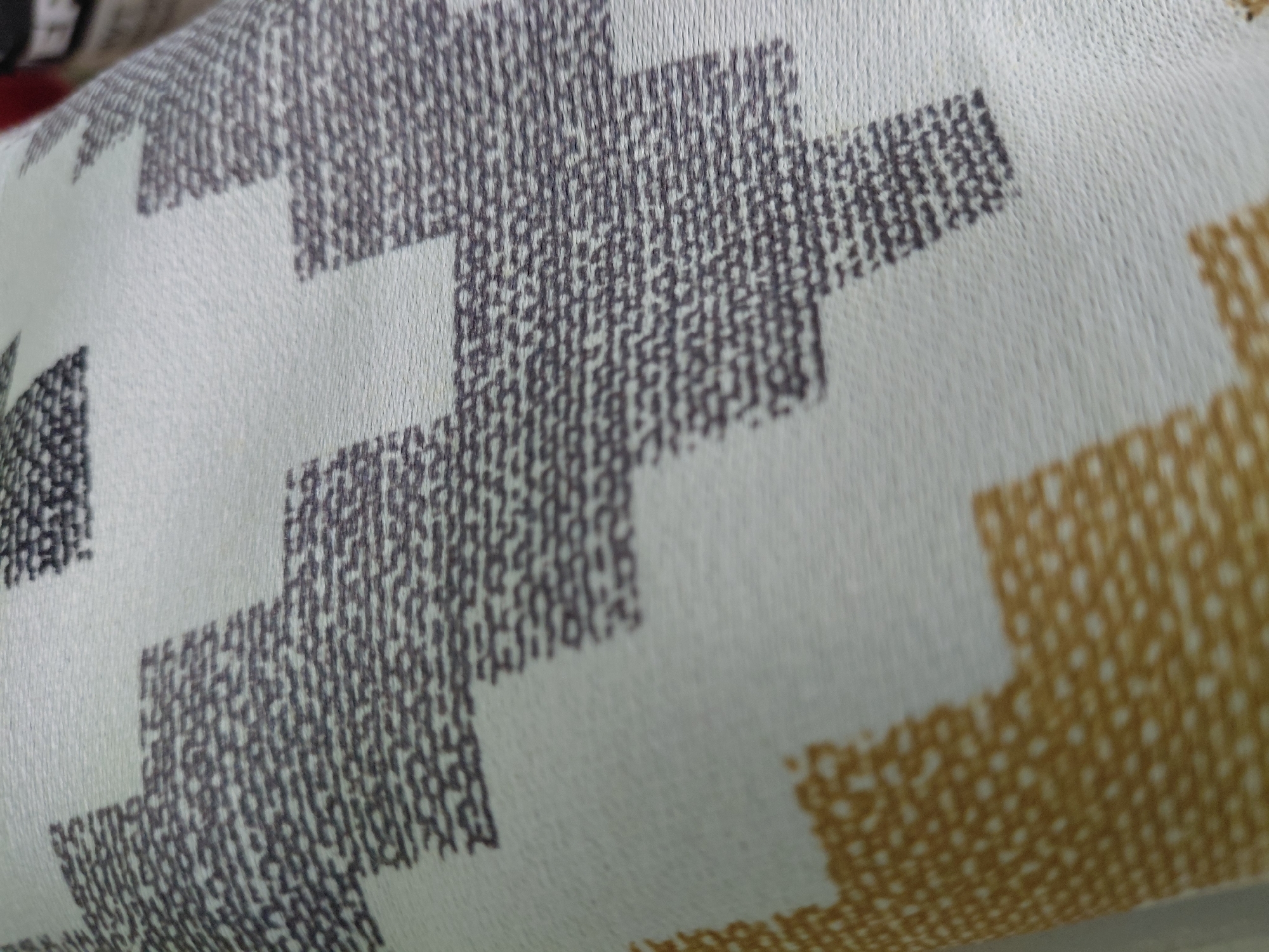 The image presents a close-up view of a textured textile or fabric, possibly part of upholstery, a blanket, or a curtain. No people are visible. The fabric features a repeating geometric pattern composed of stepped, interlocking shapes. The primary background color of the fabric is a light shade, appearing as white, off-white, or a very pale grey. The pattern elements are rendered in two distinct colors: a dark grey or charcoal and a warm mustard yellow or golden-brown.
A prominent detail is the discernible texture of the fabric. Within the colored pattern areas, the surface appears to be composed of numerous small, closely packed dots or fine stitches, suggesting a woven structure like a jacquard or a print designed to simulate such a texture. The overall surface has a subtle, tactile quality. The fabric shows a slight curve or fold, hinting that it might be draped over an object, part of a cushion, or fabric rolled up.
The lighting is soft and even, likely indoor, which effectively highlights the fabric's texture and colors without casting harsh shadows. The central portion of the image is in clear focus, with the areas closer to and further from the camera displaying a gentle blur, indicating a shallow depth of field. No text is visible in the image. The location context provided is Nadym, Russia, though there are no specific visual cues within the image itself to confirm this.