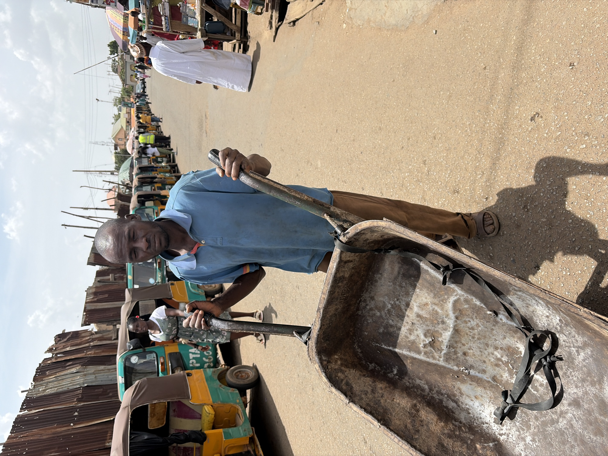 The image portrays a lively outdoor street or market scene in Gura Top, Nigeria, under bright daylight. In the foreground, a man, appearing to be of middle age, is centrally positioned. He wears a light blue short-sleeved polo shirt and brown trousers, with sandals on his feet, and has short dark hair. He firmly holds the handles of a large, heavy-duty metal cart, similar to a wheelbarrow, which dominates the lower right portion of the frame. The cart is visibly old and rusty, showing signs of extensive use, with black straps neatly tied and lying inside its basin. The man's expression is neutral and focused, suggesting he is either preparing to move or has paused during an activity involving the cart.
Behind the man, the street is made of dirt or gravel, with strong shadows indicating a sunny day, likely midday or early afternoon. A row of green and yellow "keke napep" (tricycle taxis) is lined up along the left side of the road, suggesting a common transport and market area. Adjacent to these vehicles are makeshift market stalls with corrugated metal roofs, further reinforcing the impression of a bustling local market. Several other people are visible in the background, including one individual in a white robe walking further down the street and others near the vehicles. In the distant background, power lines and indistinct buildings can be seen under a clear sky with scattered clouds. On one of the "keke napep" vehicles, the partial text "PTA 360" is discernible.