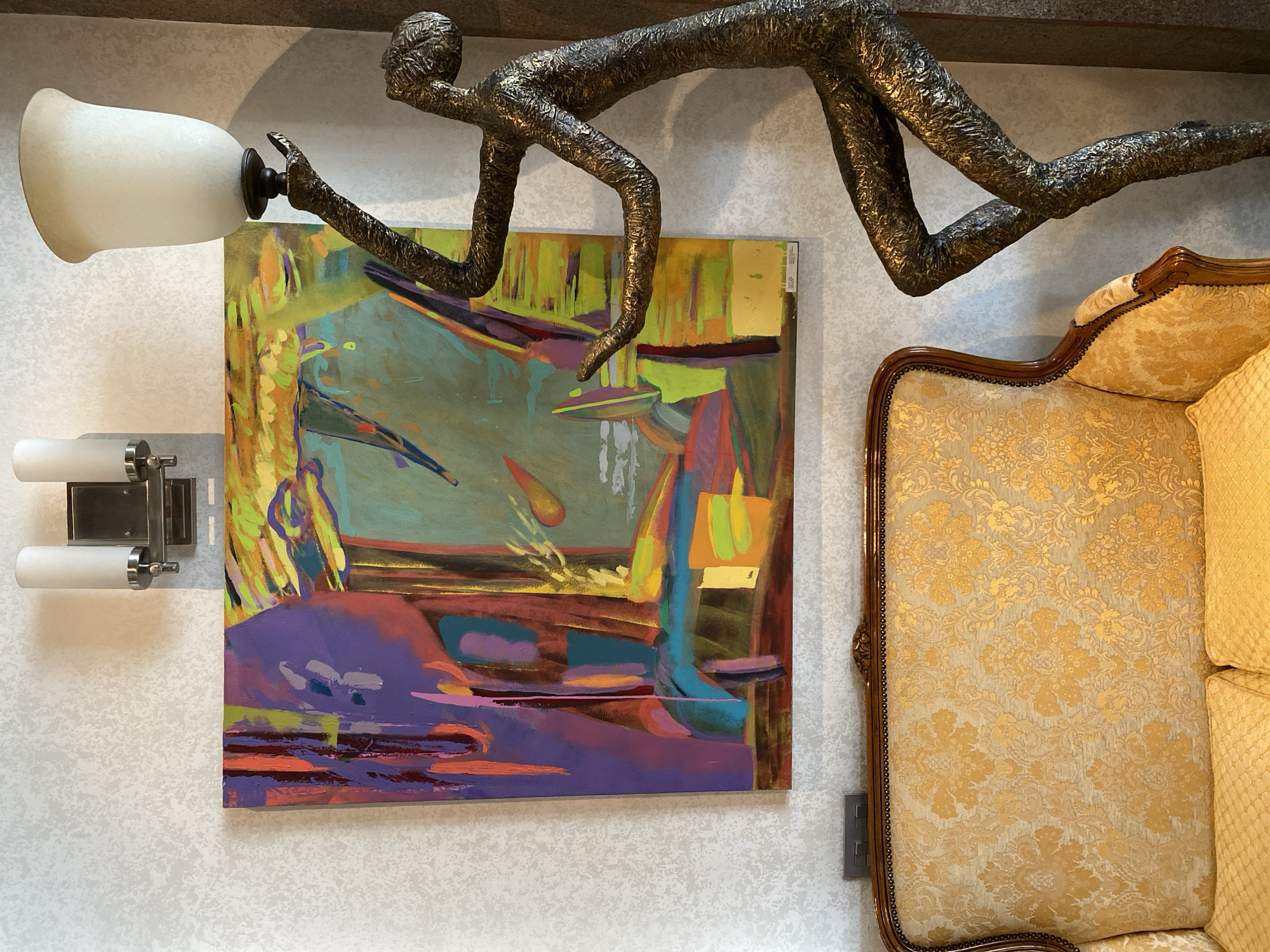 The image shows a section of an interior wall decorated with several elements. A square abstract painting with a vibrant color palette, including shades of purple, blue, green, and orange, hangs on the wall. Above the painting, a bronze-colored sculpture of a person in a crawling or reaching pose is mounted. To the left of the painting, a wall sconce with two cylindrical lights is installed. To the right, part of an ornate sofa or chair with a patterned gold fabric is visible. The wall itself has a textured, off-white wallpaper. The image is likely taken indoors, possibly in an art gallery, a hotel, or a private residence with a distinctive style. There's no concrete information given about the location being in Yekaterinburg, Russia.