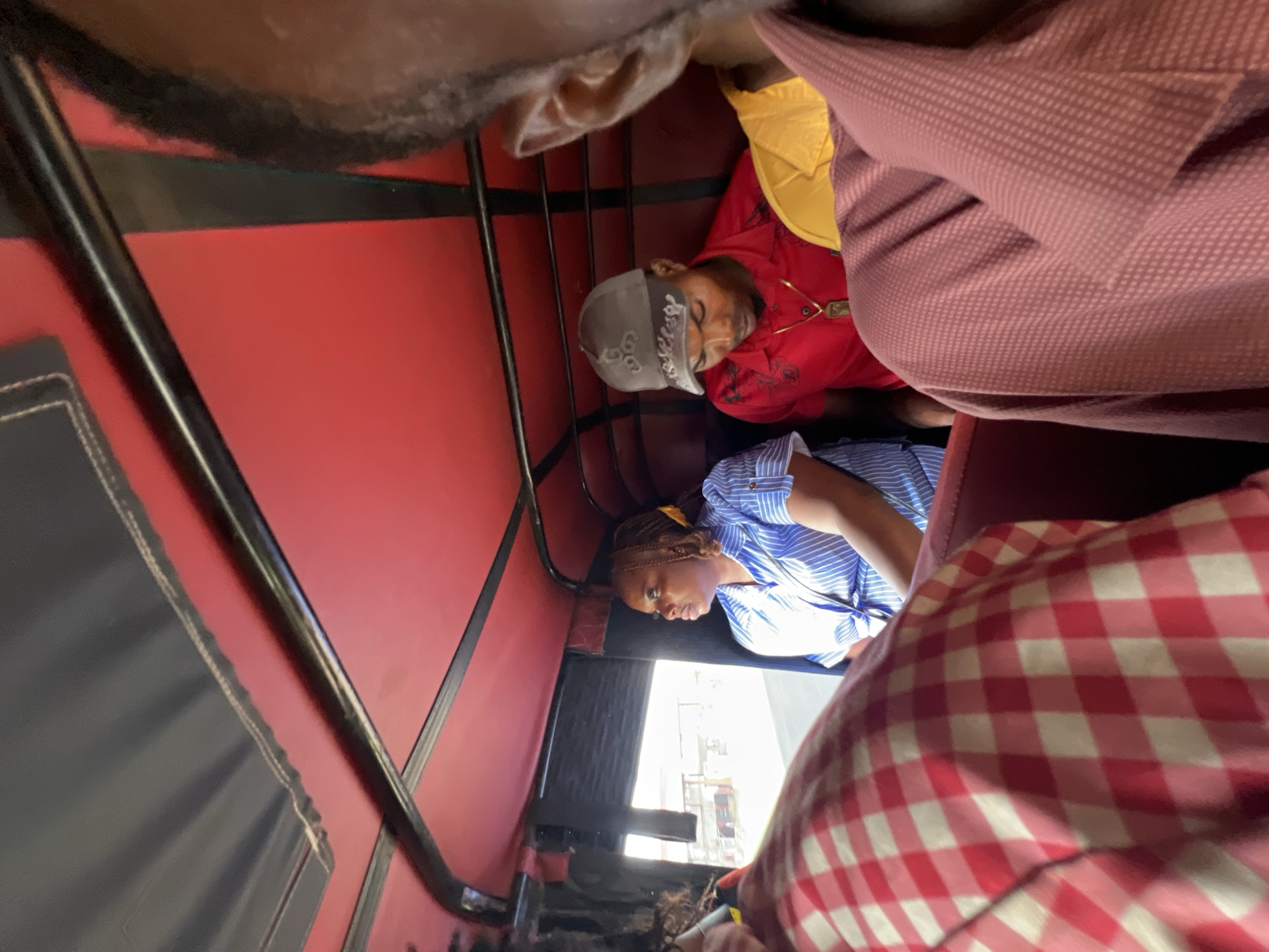 The image depicts the interior of a tuk-tuk, a three-wheeled motor vehicle commonly used as a taxi. It is captured in Nkpor, Nigeria. Four individuals are visible inside the vehicle, which has a red-colored roof lining.
In the foreground, on the left, a person is partially visible, wearing a red and white checkered shirt. To their right, a woman with braided hair and a yellow ribbon is seated, looking to the left. She is wearing a blue and white striped button-up shirt. In the middle of the vehicle, a man wearing a red polo shirt and a gray baseball cap is seated. He is looking downwards. To his right, another person is partially visible, wearing a yellow shirt. The person closest to the camera, on the right side of the frame, is wearing a maroon textured shirt. The tuk-tuk's interior appears somewhat cramped, with visible metal framing supporting the roof. Through the open side of the tuk-tuk, a blurred view of a street scene, possibly with buildings and a road, can be observed. The overall lighting suggests it is daytime.