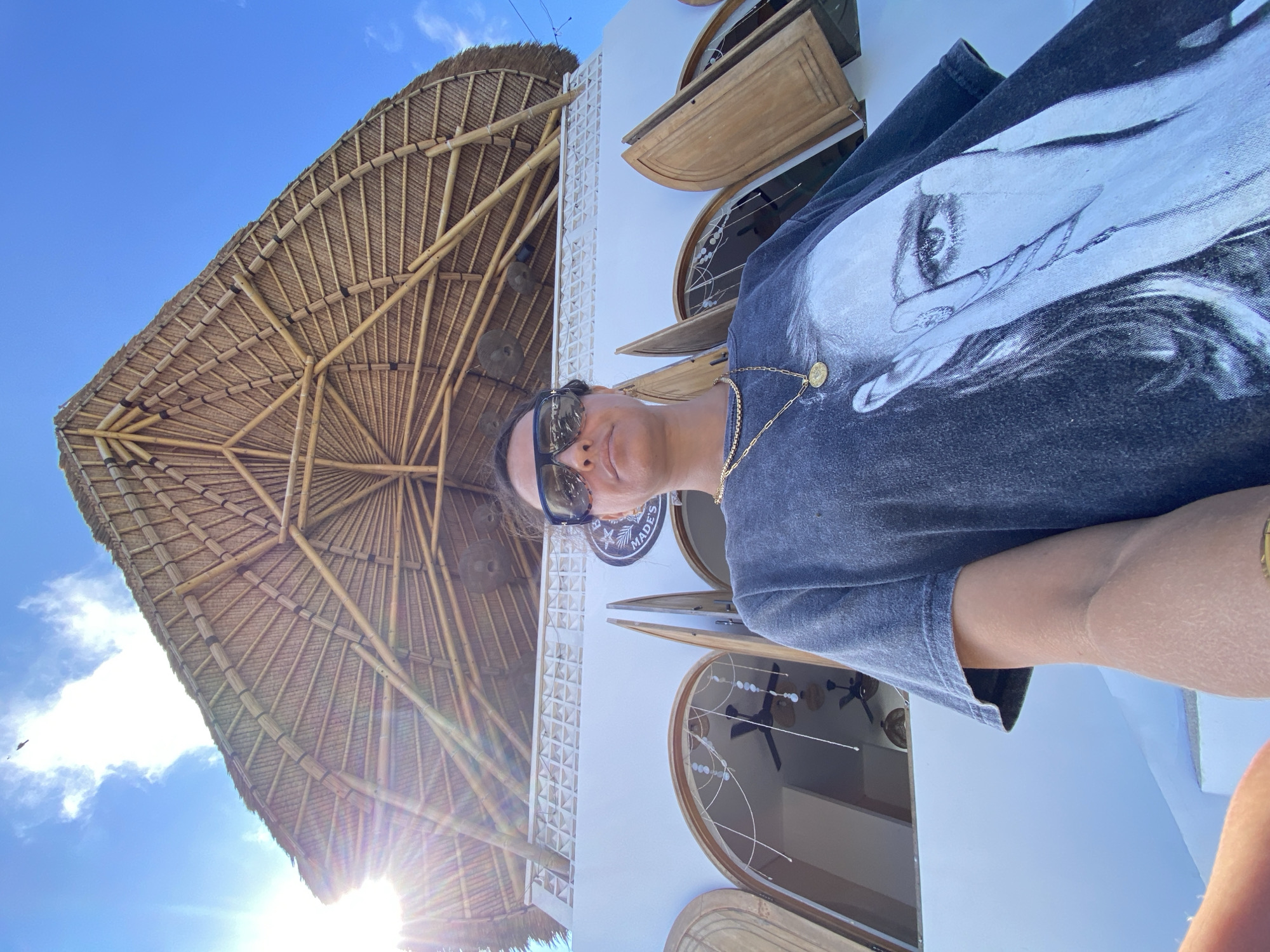 An outdoor, low-angle, wide shot captures a person standing in front of a white, two-story building with an elaborate, multi-tiered bamboo roof structure. The building features arched windows with wooden shutters on the second floor, and decorative white fretwork above them. A circular sign with a palm tree and the word "MADE'S" is visible on the facade. The person, wearing a dark gray t-shirt with a black and white graphic of a woman's face and neck, large sunglasses, and gold necklaces, is positioned in the lower right quadrant of the frame, facing the camera. Sunlight beams into the frame from the upper left, casting lens flare and illuminating the bamboo roof and the sky, which is partly cloudy. The overall impression is a tropical, possibly commercial, establishment. The sign indicates the location as "MADE'S" in Kuta Selatan, Indonesia.