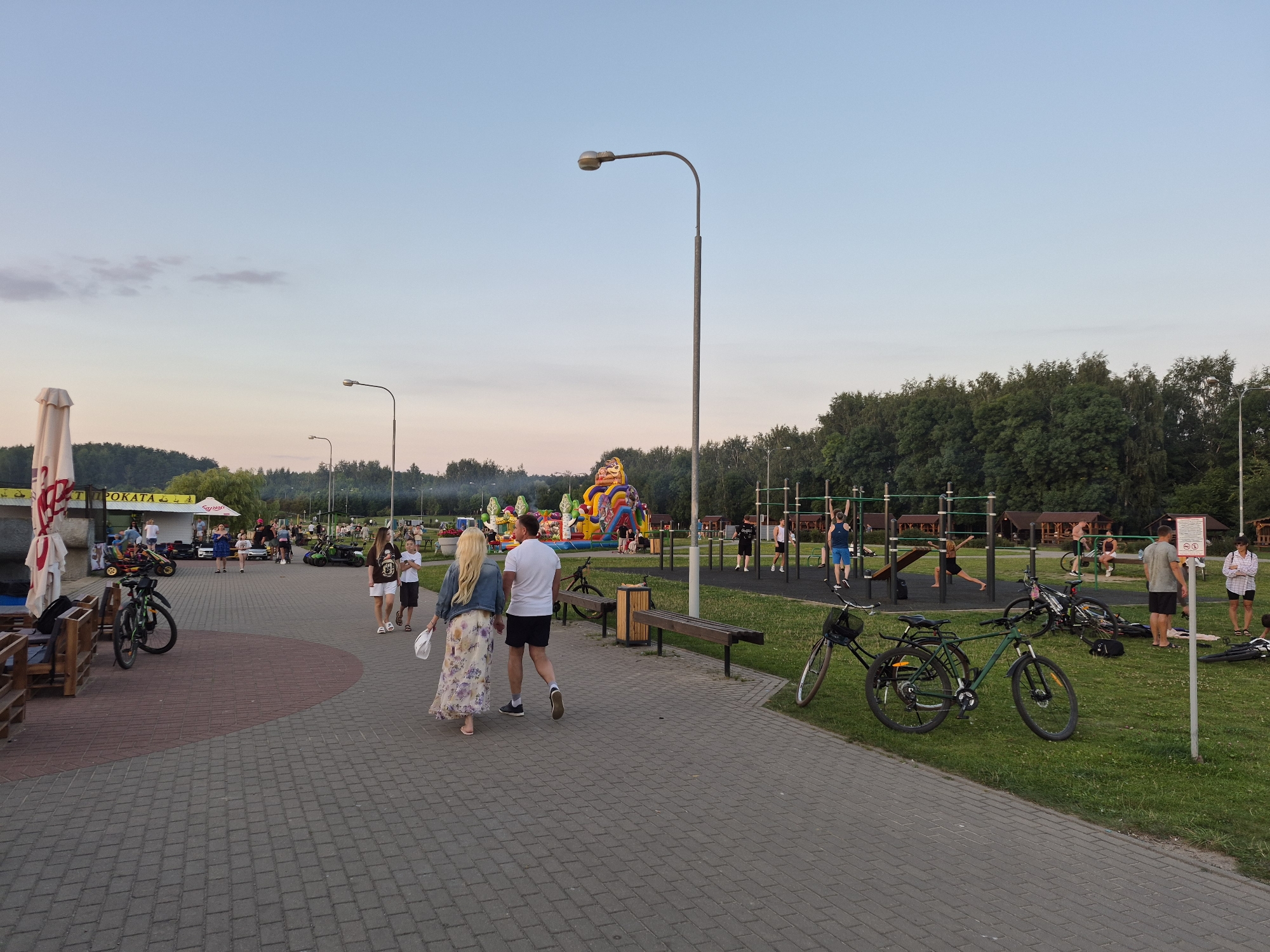 The image depicts an outdoor recreational area in Minsk, Belarus, during dusk. A paved walkway, made of interlocking bricks, dominates the foreground. Several people are walking or standing in the scene. A couple, a woman in a floral dress and a man in a white t-shirt and black shorts, walks away from the camera on the walkway. To their left, a large inflatable playground structure, possibly a bouncy castle, is visible with several children playing on it. Further in the background, there is a children's amusement area with go-karts. A dedicated area with exercise equipment, likely an outdoor gym, is situated on the right side of the image, with several individuals using the equipment. Bicycles are parked on the grass and along the walkway. Trees line the background, and a pale sky suggests the late evening hours. The overall ambiance is that of a public park or leisure complex.