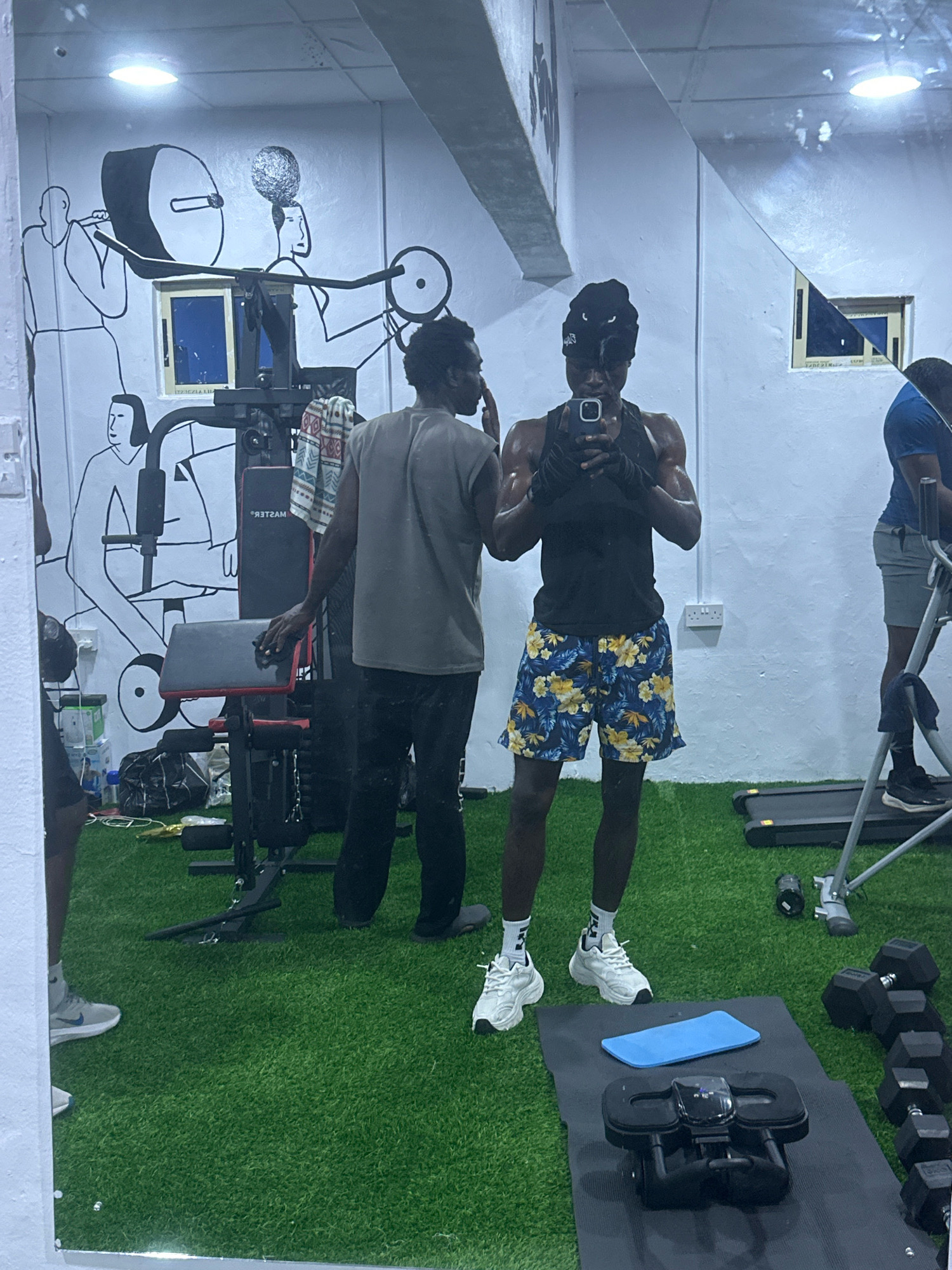 In a gym located in Badagry, Nigeria, a young Black man stands in front of a mirror, taking a selfie with his phone. He is wearing a black tank top, a black beanie, and patterned shorts. His arms are muscular, and he appears to be sweating. In the mirror's reflection, another Black man in a grey t-shirt and black pants is visible, standing near a weight machine. The gym has artificial turf flooring and a mural of stylized figures lifting weights on the wall. Other gym equipment, including dumbbells and a treadmill, are also present.