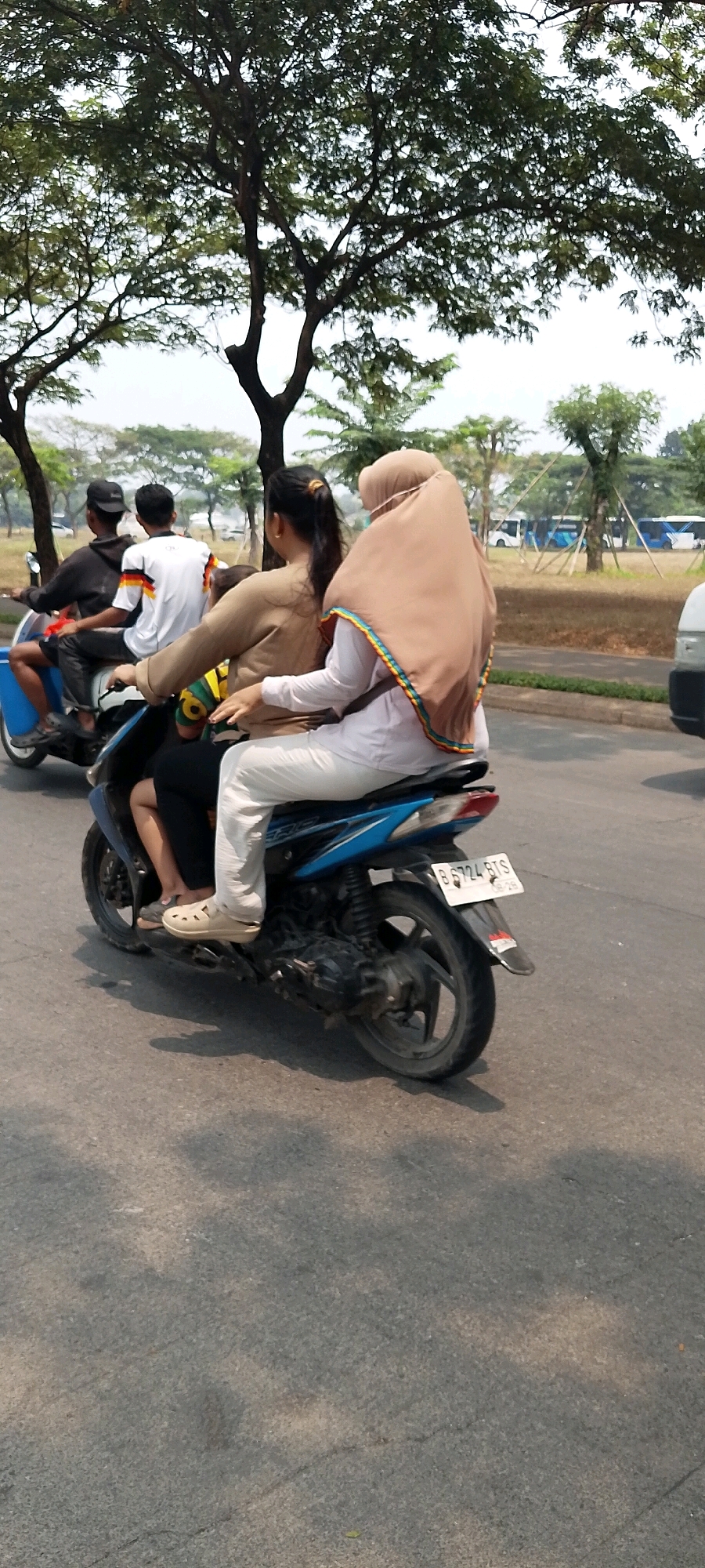 The image depicts a street scene in Bekasi, Indonesia, with a focus on a blue and black motorcycle carrying three people. The motorcycle is in motion on an asphalt road, casting shadows from overhead trees. The people on the motorcycle are dressed casually. The rider in the back, wearing a light brown hijab and white pants, appears to be a woman. In front of her, another woman wearing a tan sweatshirt and dark pants is seated. A third person, partially obscured, is seated in front of them. In the background, several trees line the side of the road, and a parking lot with buses is visible. The license plate on the motorcycle reads "B 6724 BXS". The overall atmosphere suggests a typical urban street environment in Indonesia.
