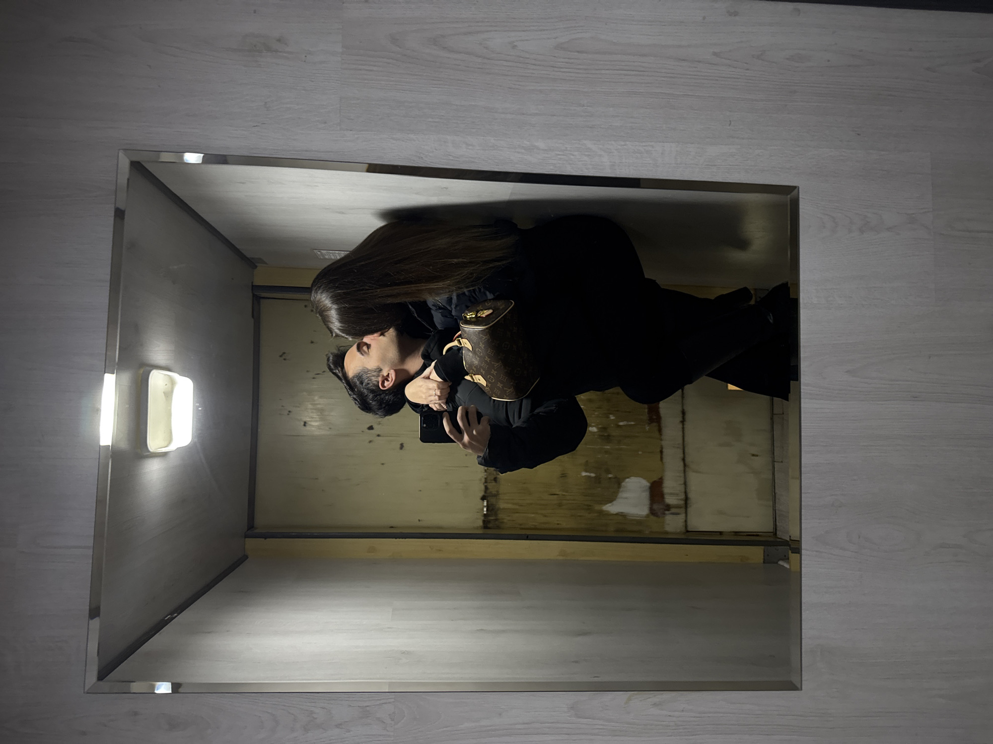 A couple is reflected in a square mirror. The woman has long, brown hair and is wearing black. The man has dark hair and a beard, and is also wearing black. He is holding a cell phone. The reflection shows a shabby interior with bare walls and wooden supports. The environment outside the mirror has white wooden planks.
Without additional data, it is impossible to pinpoint the location in Sofia, Bulgaria.