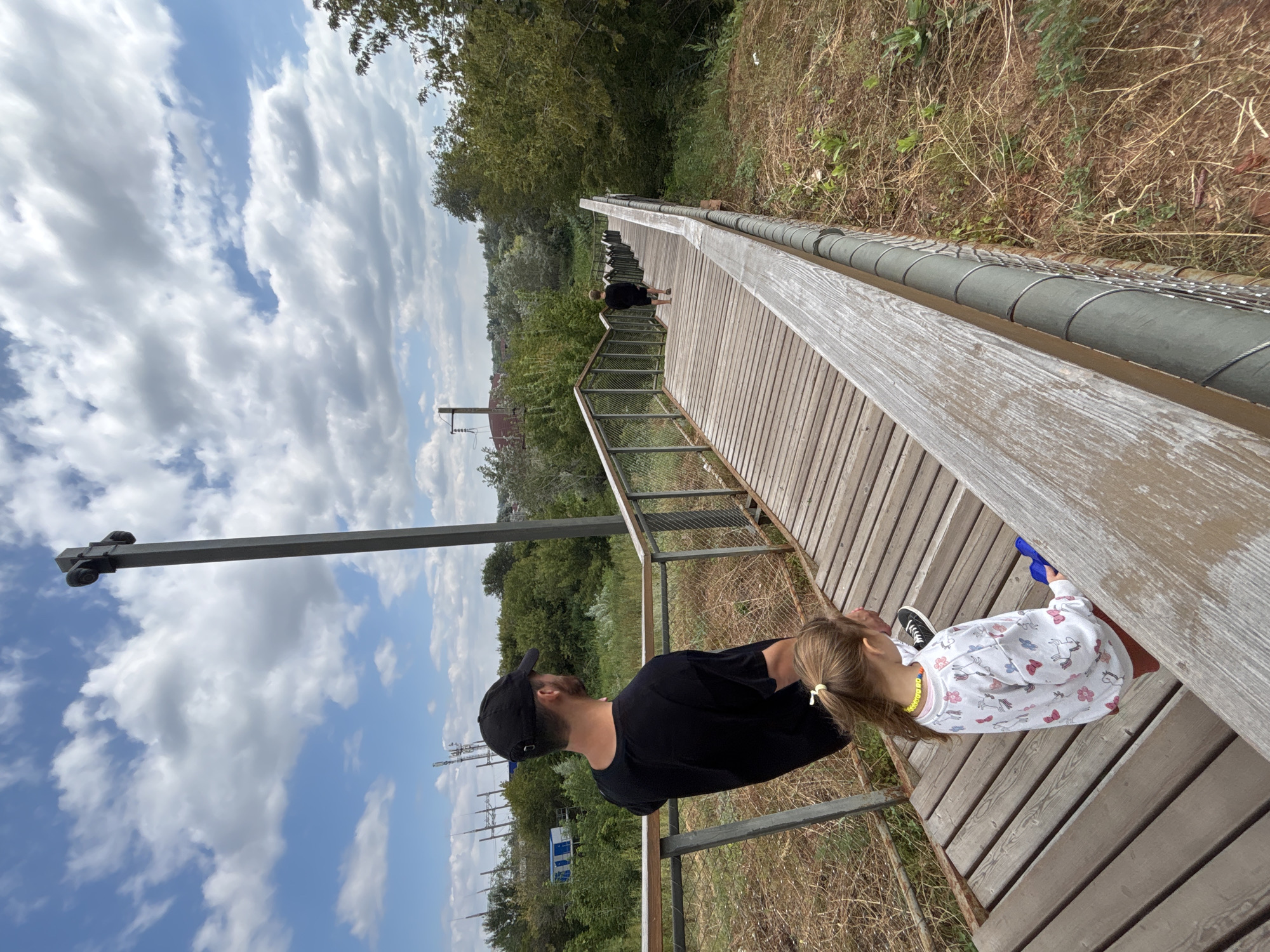 The image captures an outdoor scene with a wooden staircase winding down a grassy embankment under a partly cloudy sky. In the foreground, a man wearing a black t-shirt and baseball cap stands looking away from the camera, with his back to the viewer. A young girl, wearing a white sweatshirt with colorful animal prints and red shorts, stands on the staircase in front of him, holding a blue object. Further down the staircase, a child in a black shirt and shorts walks away from the camera. The staircase is bordered by metal railings and a rough, natural landscape of dry grass and greenery. In the background, trees and distant structures, including power lines and industrial elements, are visible, suggesting an urban or semi-urban environment. The overall impression is of a recreational area, possibly a park or viewpoint, located in Orenburg, Russia.