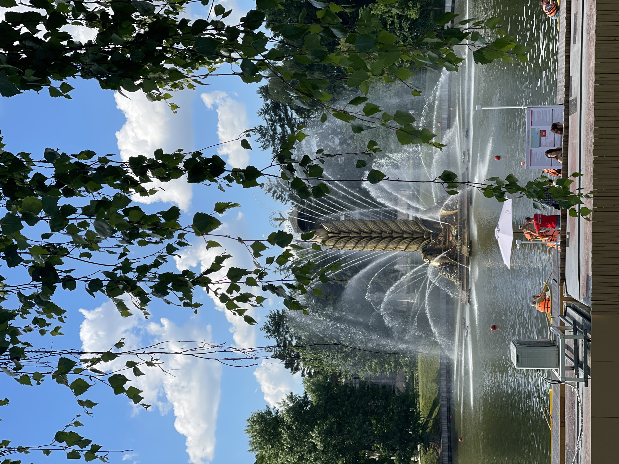 The image depicts a vibrant outdoor scene on a bright, sunny day, centered around a grand, ornate fountain in a large body of water, likely a pond or lake. The fountain, appearing golden or bronze, features an elaborate design with multiple jets of water spraying high into the air, creating a misty halo around its structure.
On the right side of the image, a wooden boardwalk or dock borders the water. Several people are visible here; some are seated under a white umbrella, while others are in small yellow boats or rafts on the water, all wearing orange life vests. A large white information sign stands prominently on the bank, next to which a grey outdoor unit is mounted on a metal frame. Red buoys float in the water.
Lush green tree branches and leaves frame the top and left sides of the scene, with more dense foliage visible on the far bank of the water body. The sky is clear and blue, dotted with white, fluffy clouds, suggesting pleasant weather.
The activity taking place appears to be leisure and recreation. People are enjoying the lakeside environment, potentially engaging in boating or simply relaxing by the water.
Notable details include the intricate design of the fountain and the clear, bright daylight. Visible text on the white information sign includes "ВОДНАЯ СТАНЦИЯ" (Water Station), "ПРАВИЛА ИСПОЛЬЗОВАНИЯ" (Rules of Use), "ПРОКАТ ЛОДОК" (Boat Rental), and "ПРОКАТ КАТАМАРАНОВ" (Catamaran Rental), indicating this is a public recreational area with boat rental services. There also appear to be some Cyrillic characters like "ОРНАТ" and "КОМ" on the fountain's golden structure.