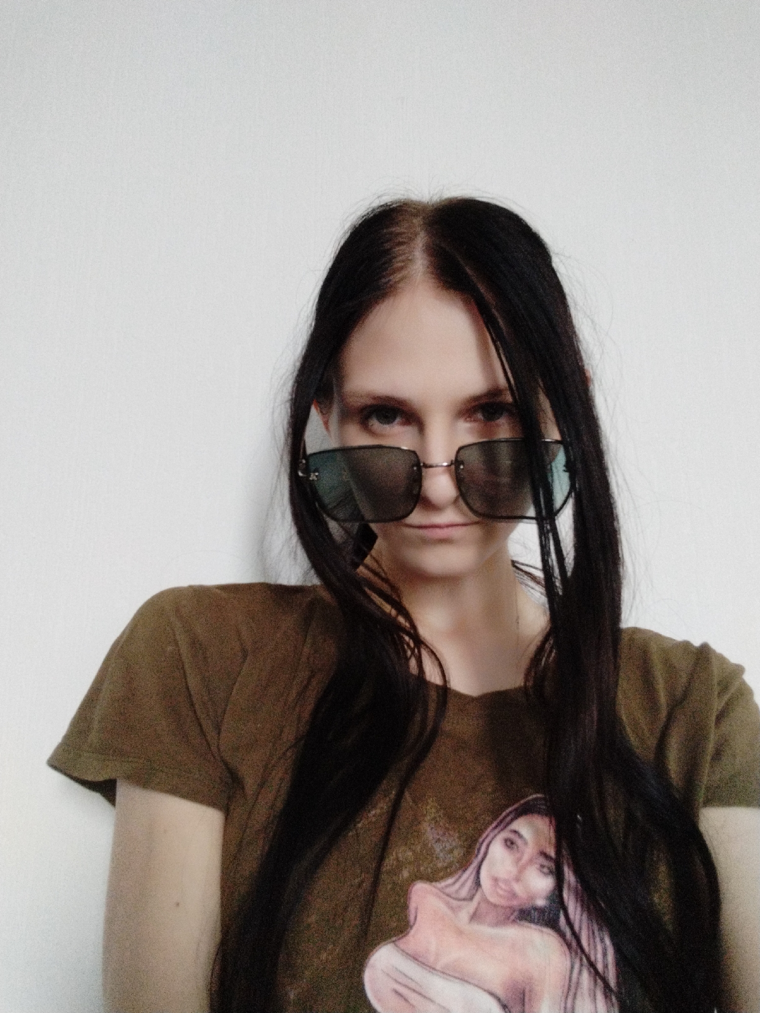 A young woman with long, dark hair and fair skin, wearing a dark olive green t-shirt with a graphic print of a woman, and large, dark-lensed sunglasses, looks directly at the camera with a neutral expression against a plain white background. Her hair is partially obscuring her face, framing her features. The lighting is flat and even, suggesting an indoor setting with artificial light.