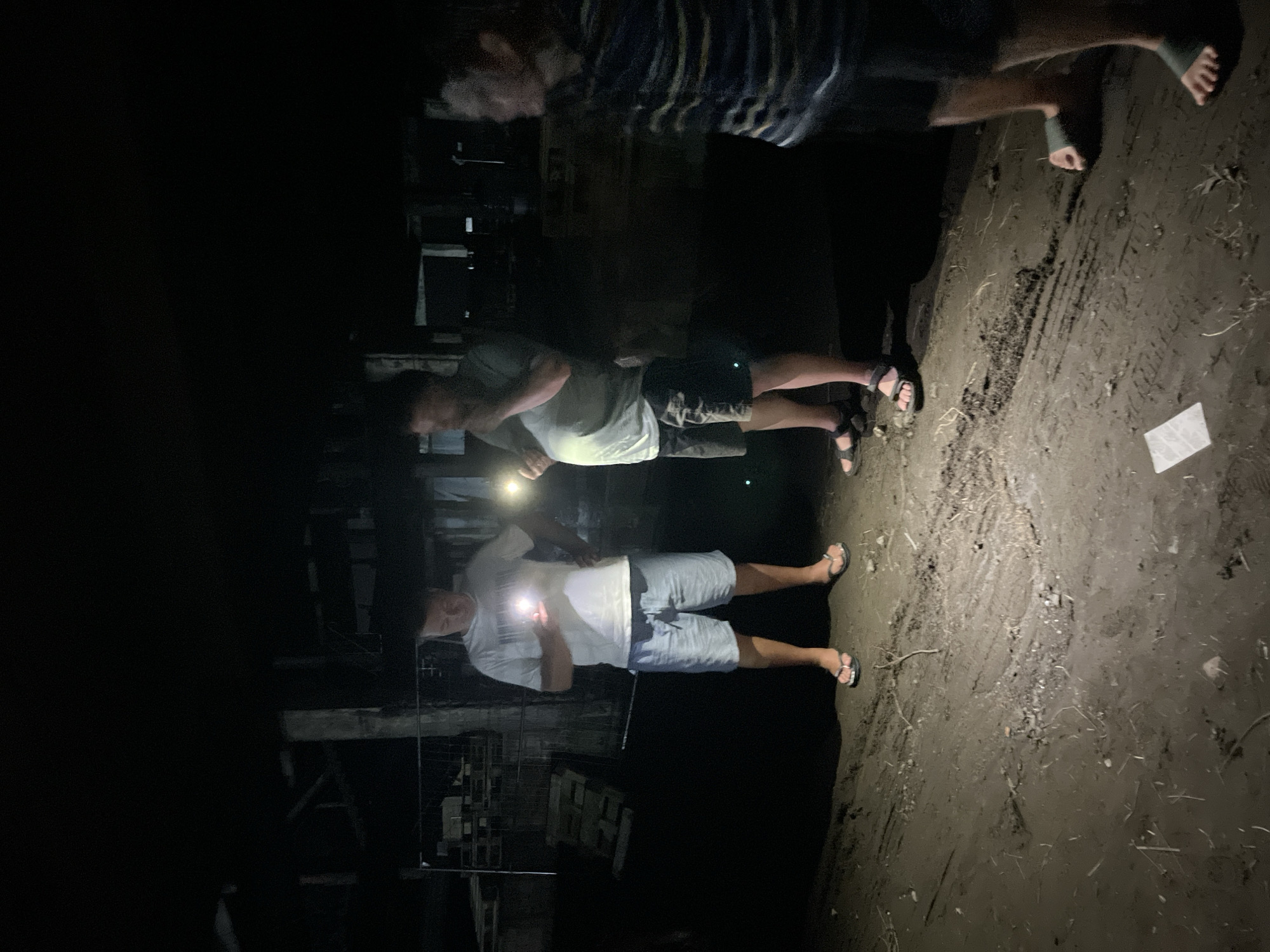 Three individuals, two adult males and one child, stand in a dimly lit outdoor environment that appears to be a construction site or a derelict area, possibly in Poti, Georgia. The ground is composed of dirt and debris, with visible tire tracks. In the background, scaffolding and building structures are obscured by darkness. The individual on the left, an adult male wearing a white t-shirt and light-colored shorts, is holding a light source, illuminating his surroundings. The central figure, also an adult male, is wearing a light green t-shirt and dark shorts, and is also holding a light. The third individual, a child on the right wearing a striped shirt, is partially visible and appears to be looking away from the camera. The overall atmosphere is one of an impromptu gathering or exploration in a dark, unrefined setting.