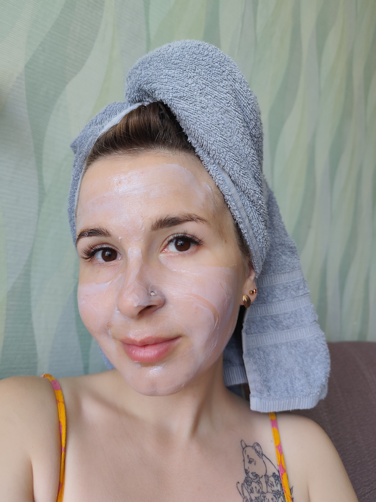 A woman with a towel wrapped around her head is facing the camera. She has a white facial mask on her face. The woman is wearing a yellow and pink tank top, and a tattoo is visible on her left chest. The background is a pale green wallpaper with a wavy design. The location is not provided in the image.