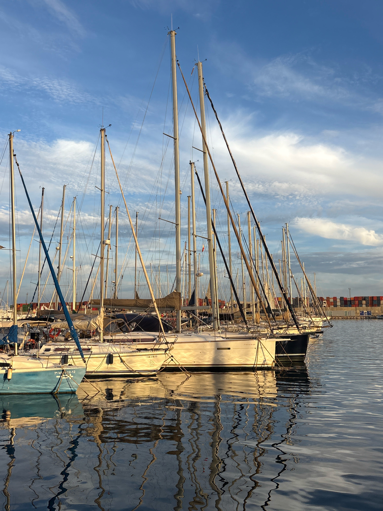 This is a photograph taken at sunset in the Marina Real Juan Carlos I in Valencia, Spain. The foreground is dominated by the reflective surface of the water, mirroring the masts and rigging of several docked sailboats. The boats are mostly white with some darker hulls, and their reflections create intricate patterns on the water.
In the mid-ground, a line of sailboats is moored side-by-side. The masts of these sailboats reach towards the sky, intersecting with the wispy clouds in the blue sky. The sky itself is a mix of blues and whites, with the setting sun casting a warm, golden light on the scene.
In the background, a modern industrial area with stacked shipping containers can be seen, contrasting with the natural beauty of the marina and the sky. A few people are visible on the docks, but they are small and indistinct. The overall impression is one of tranquility and the end of a day at sea.