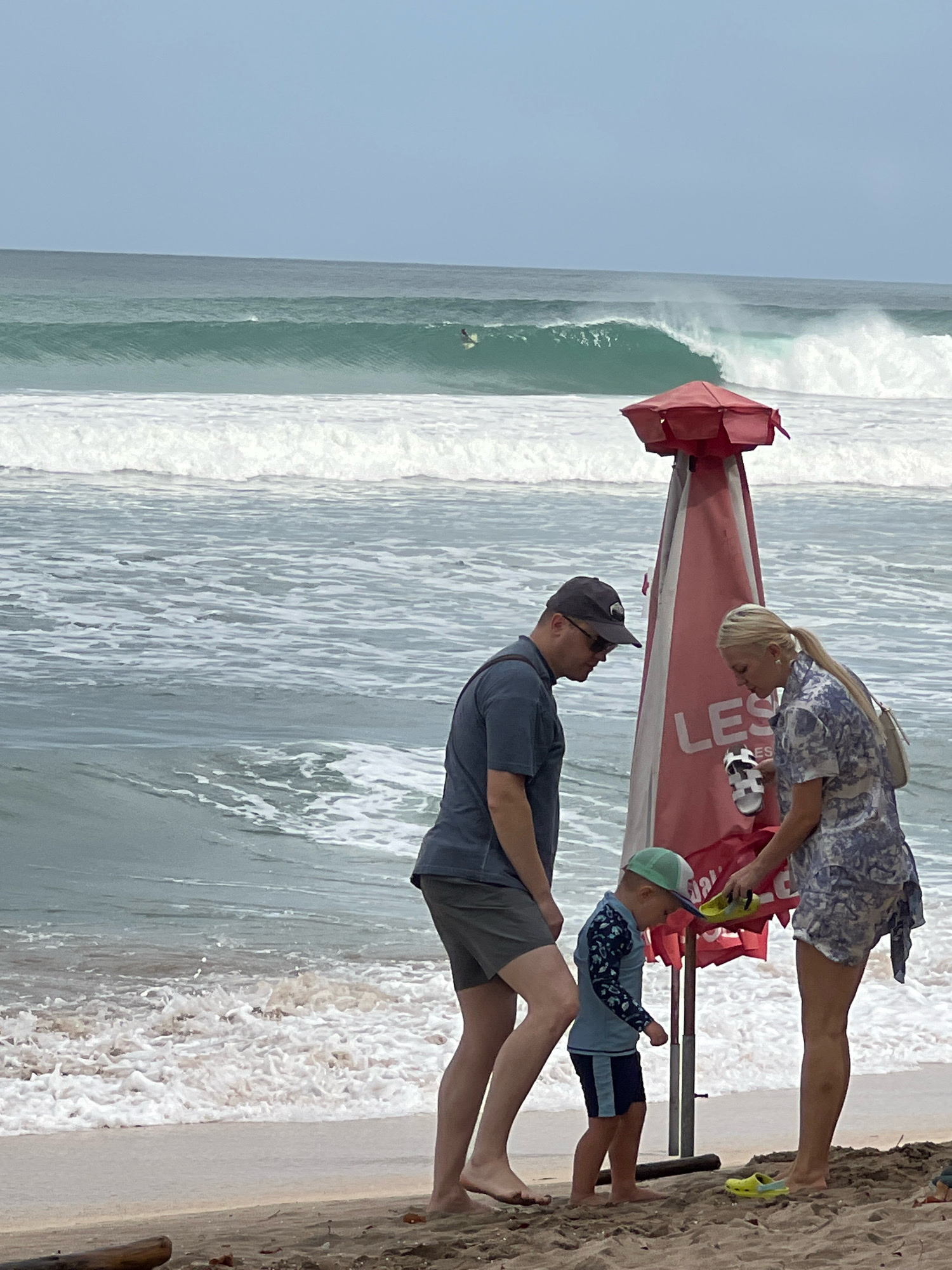 A family stands on the beach in Kuta, Indonesia, near a red lifeguard umbrella. The ocean behind them has large waves, with a surfer visible in the distance. A man, woman, and young boy are the subjects. The man is wearing a dark t-shirt, grey shorts, and a baseball cap. The woman is wearing a patterned dress and carrying a white purse. The boy is wearing a blue rash guard, blue shorts, and a green hat. They appear to be interacting with items near the lifeguard umbrella. Yellow crocs are visible on the sand near the woman and the boy.