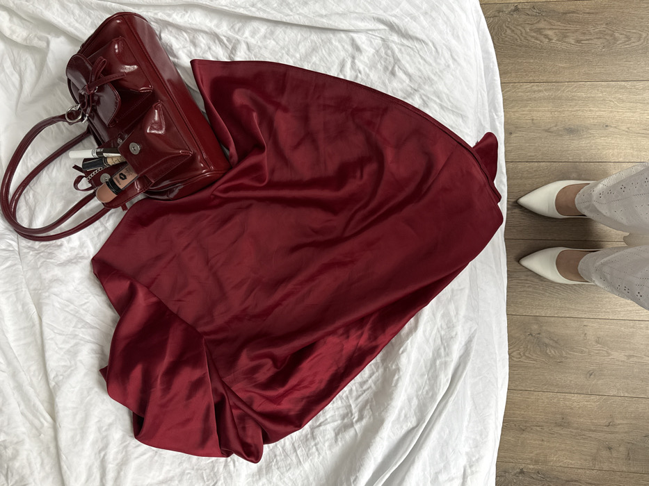 An overhead view depicts an indoor scene with a person's feet positioned on a light brown wooden floor, looking down towards a collection of items arranged on a white, wrinkled fabric surface. The specific city location of Krasnodar, Russia, cannot be determined from the visual evidence within the image.
The upper portion of the image shows a large piece of deep red, satiny fabric, possibly a skirt or dress, draped across the white fabric, exhibiting various folds and creases. To its right, a glossy, dark red handbag with two handles is placed on the white fabric. The handbag features multiple compartments, some adorned with small bows. An open front pocket of the handbag reveals several cosmetic items, including tubes of lip gloss or foundation, one marked with a red lips graphic, and other cylindrical makeup products.
In the lower section of the image, two feet are visible, clad in white, pointed-toe, low-heeled shoes. The individual is also wearing white trousers or pants with a textured, possibly embroidered or eyelet, pattern, the lower portion of which is visible above the shoes. The wooden floor boards extend from the bottom of the frame to where they meet the white fabric surface, creating a clear horizontal division in the scene. The lighting is even and soft, suggesting an indoor domestic setting. No other individuals or active interactions are observed.