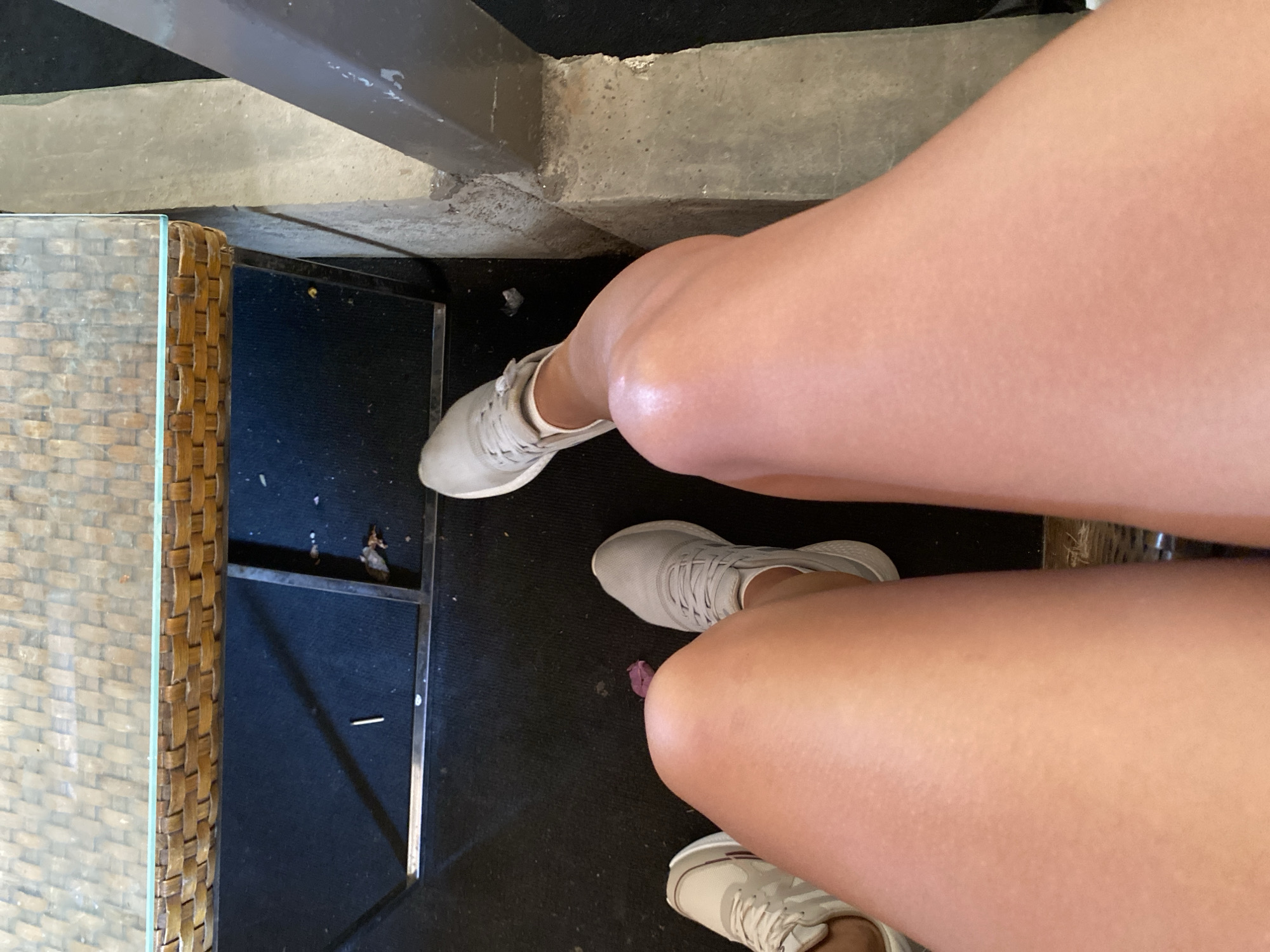 The image shows a close-up view of a person's legs, wearing casual shoes. The perspective is from above, looking down at the knees and thighs. The legs are tanned and appear to be illuminated by sunlight, highlighting the texture of the skin. The person is wearing light-colored athletic shoes. In the background, a woven table is visible, with a dark carpeted floor beneath it. There are some small debris on the floor, including a thin metal object and some organic matter. The scene appears to be an outdoor or semi-outdoor setting, possibly a cafe or patio. There is no indication of the location being in Aksu, Turkey.