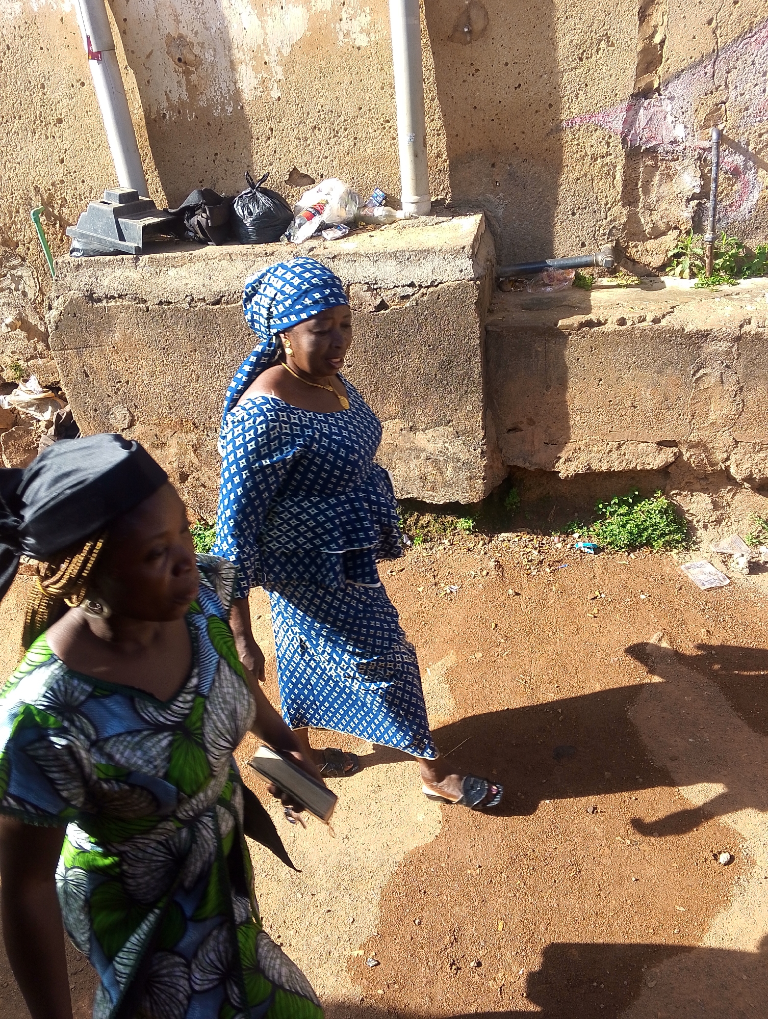 Two women walk on a dirt path in Jos, Nigeria. The woman in the foreground wears a green and blue patterned dress and a black head covering. She is carrying a small, rectangular object. The woman behind her wears a blue and white patterned outfit and headscarf. Behind them is a weathered concrete wall with pipes running along it, and some trash on top. The ground is reddish-brown dirt. Shadows are cast across the ground.