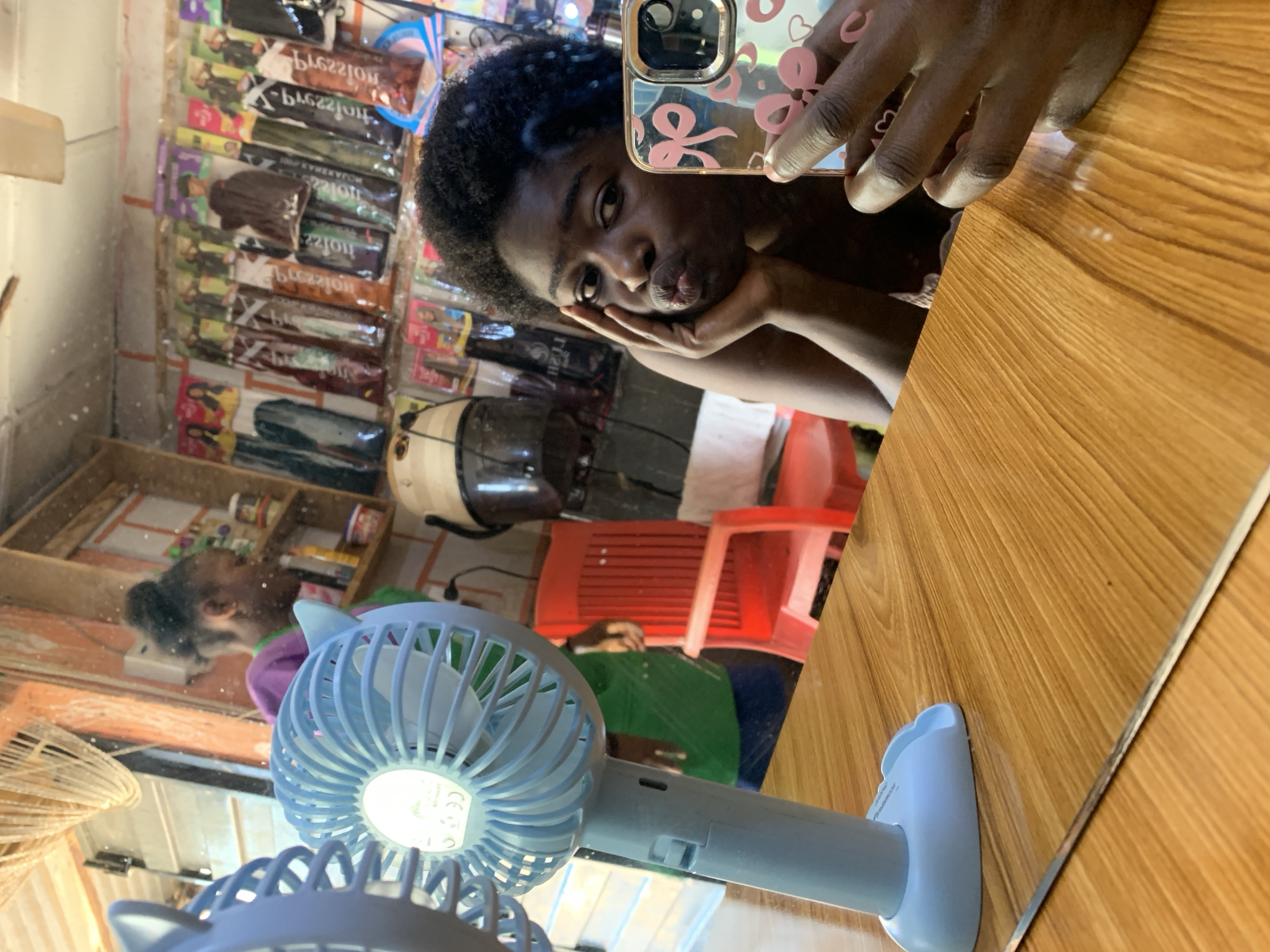 A mirror selfie taken inside a hair salon in Rafin Par, Nigeria. A young Black woman is holding a cell phone with a pink case in her right hand and resting her chin on her left hand. Her face is turned towards the mirror, and she has dark, curly hair. The background of the mirror shows salon items such as hair extensions, a hair dryer, and a shelf with beauty products. There are other people present in the salon. A small blue fan is positioned on the table in front of the mirror. The surface the fan is resting on has a light brown wood grain pattern.