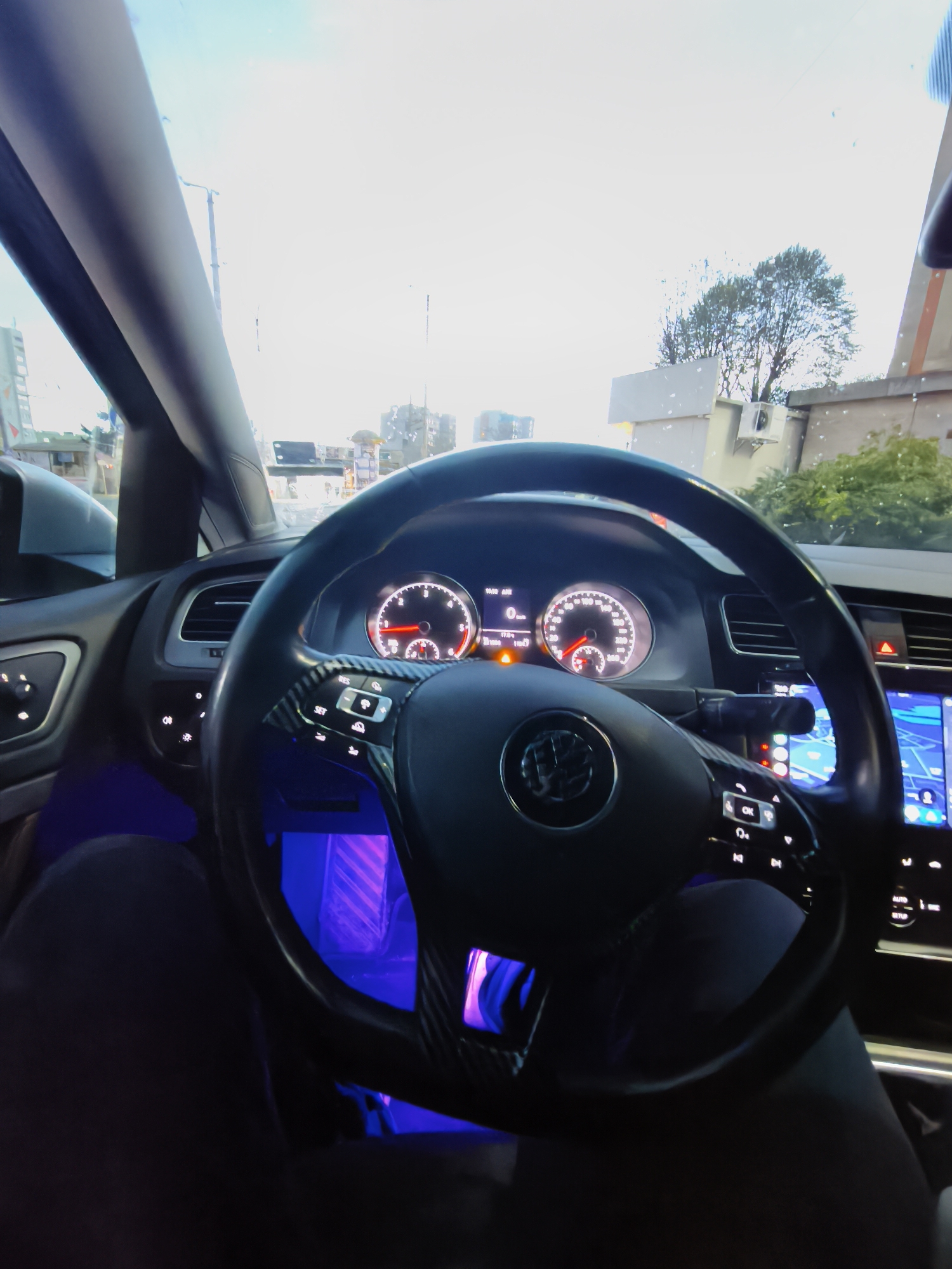 This media file captures a first-person perspective from the driver's seat of a modern car, looking out through the windshield. The vehicle's interior is dark, contrasted by various illuminated elements. The most prominent feature is the steering wheel, bearing a Volkswagen logo and adorned with carbon fiber-style accents. It has multiple control buttons, including "RES" and "SET," for cruise control and media functions.
Behind the steering wheel, the instrument cluster is brightly lit. The speedometer clearly indicates "0 km/h," suggesting the car is stationary or moving very slowly. A digital readout shows "10:30 AM" and a temperature of "11.0°C." Several warning lights are active on the dashboard, including an orange indicator and a red triangular warning symbol, possibly indicating a system alert or an unfastened seatbelt. To the right, a large infotainment screen displays what appears to be a navigation map with a blue interface. Blue ambient lighting illuminates the driver's footwell area, casting a distinct glow on the lower dashboard and the driver's legs, which are visible clad in dark trousers.
Through the windshield, a bright, somewhat hazy daytime sky dominates the upper portion of the view. The glass has visible water droplets or smudges. In the background, typical urban structures like buildings, a utility pole, and trees are discernible. The setting appears to be an urban or suburban area, consistent with the provided location context of Burgas, Bulgaria. The combination of bright exterior light and illuminated interior features creates a dynamic visual.