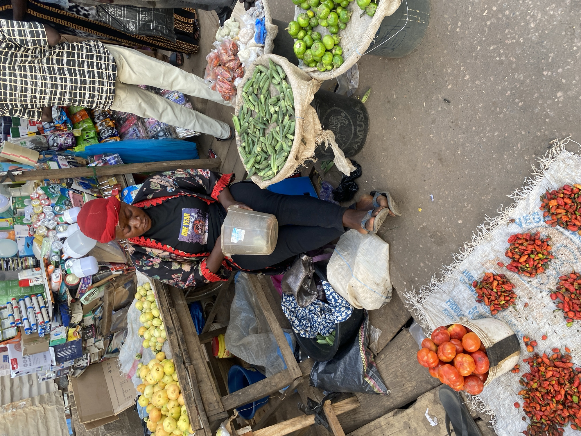 The image portrays an outdoor market stall in Sabon Tasha, Nigeria. A dark-complexioned female is seated on a low wooden structure, positioned centrally in the foreground. She wears a red headscarf, a black t-shirt with "ONE YEAR..." text, a black floral long-sleeved top with red trim, black trousers, and blue flip-flops. Her gaze is directed downwards towards a translucent plastic cup she holds. To her immediate right, the lower torso and legs of a male are visible, clad in light-colored pants and a black-and-white checkered shirt, standing.
The market stall features a variety of produce and goods. On a wooden table to the left, two distinct piles of light yellow, roundish fruits or vegetables are displayed on a white fabric. At ground level, a large dark bucket, partially covered with woven fabric, is filled with green okra. Adjacent to it, another large dark bucket, also partially covered, contains green, roundish fruits or vegetables. In the bottom foreground, a white woven sack is spread on the ground, holding a white bucket of red tomatoes and several scattered piles of small, red and orange chili peppers.
The background consists of a densely packed market stall structure, appearing to be constructed from corrugated metal and wood. This stall is laden with numerous packaged items, including various lanterns, batteries, and other small household or electronic goods. Miscellaneous objects such as black plastic bags, a blue plastic bucket, and a yellow container are on the ground around the seated woman. The ground surface is an uneven, worn reddish-brown.