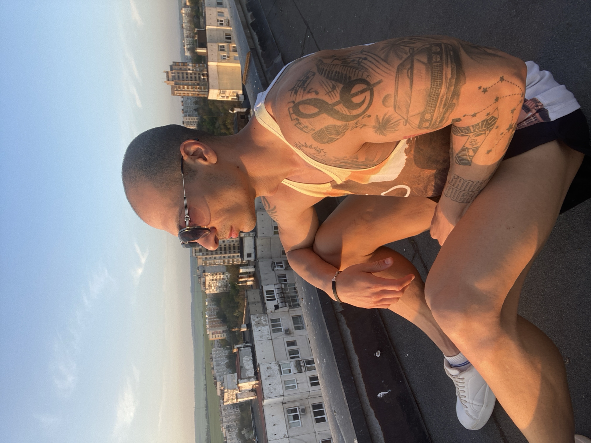 A man with short, dark hair and sunglasses is seated on a flat, dark rooftop, looking downwards and to his left. He wears a white tank top with an abstract pattern, black shorts, and white sneakers with white socks featuring blue stripes. His left wrist has a simple black bracelet.
The scene is illuminated by warm, golden light, suggesting either sunrise or sunset, with prominent shadows cast on his body and the rooftop. His arms and shoulders are heavily tattooed. On his left arm and shoulder, designs include a large looping symbol (possibly an infinity sign), musical notes, a microphone, palm trees, and what appears to be a detailed drawing of a car or boombox. His right forearm features a tattoo of an Egyptian-style pharaoh's head, a constellation of stars connected by lines, and the distinct text "GAME OVER."
The background reveals a sprawling urban landscape, characteristic of Pleven, Bulgaria, seen from a high vantage point. Numerous multi-story residential buildings fill the middle ground, while in the far distance, a flat, green agricultural field stretches to the hazy horizon. The sky is clear blue with a few faint, wispy clouds. The man's posture appears reflective or relaxed as he observes the view.
